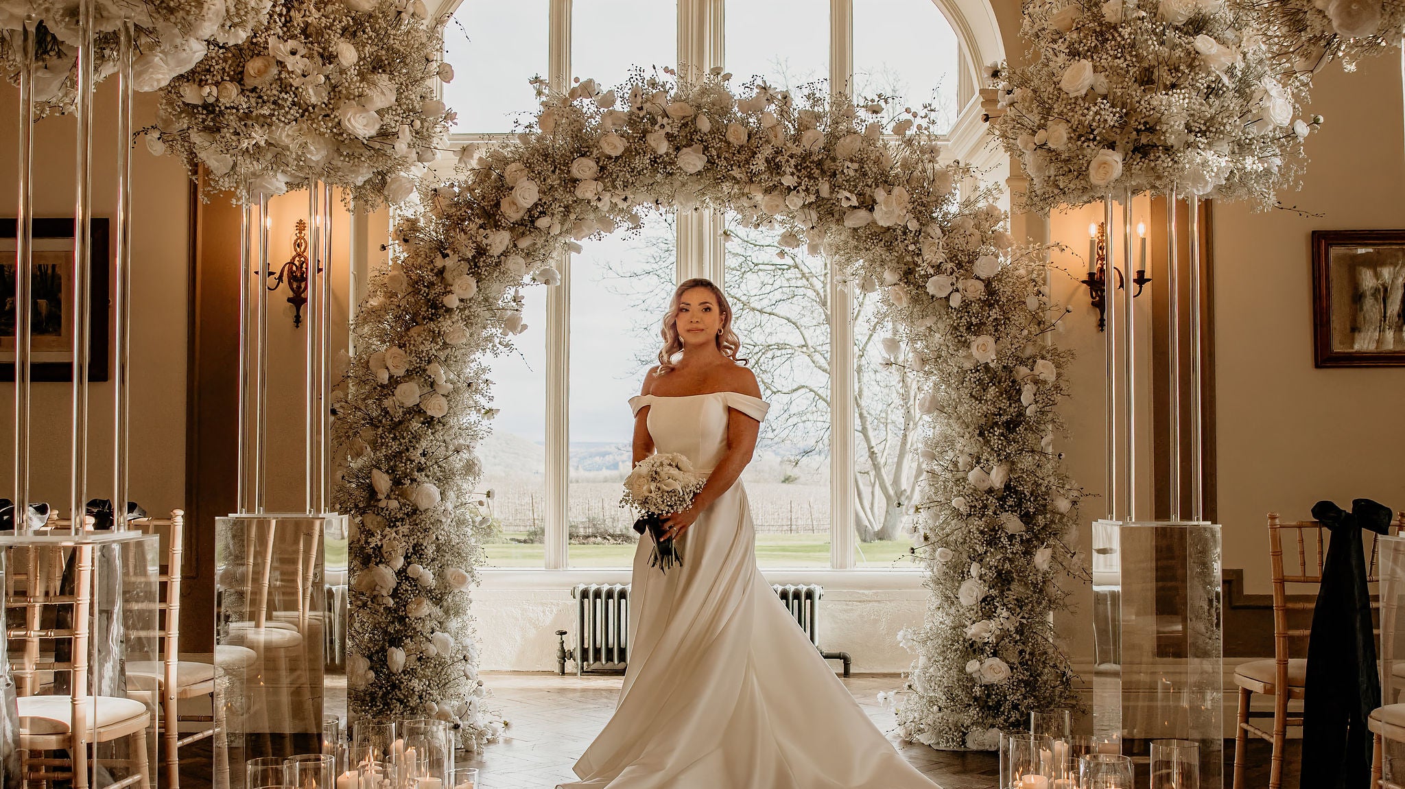 Black and white wedding arch, with bridge holding a white bouquet in a large statley home in front of a large indown, with candles down the isle