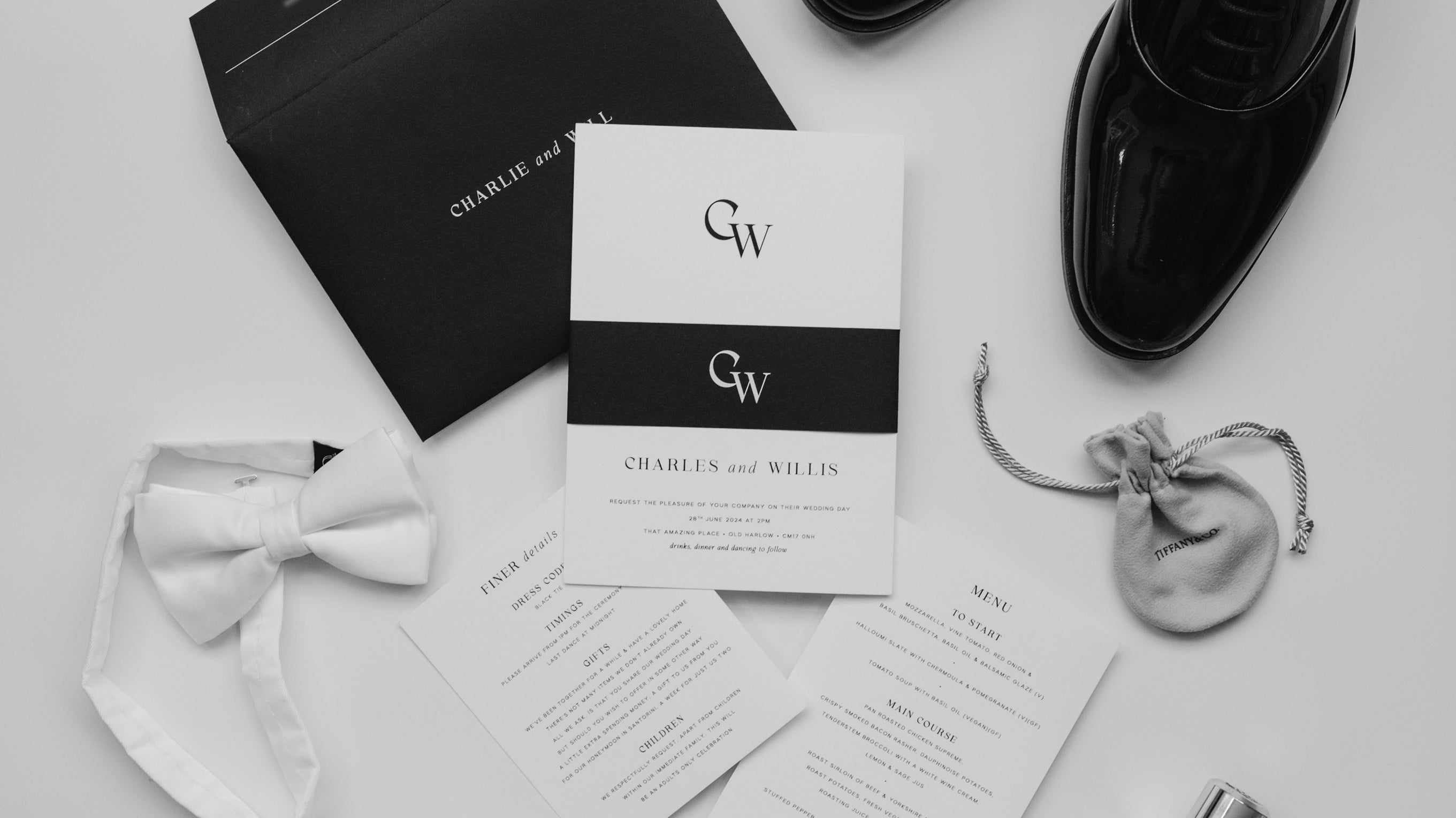 Black and white wedding invite suite, pcitured as a falt lay with black belly band, grooms shoes, aftershave and wedding rings