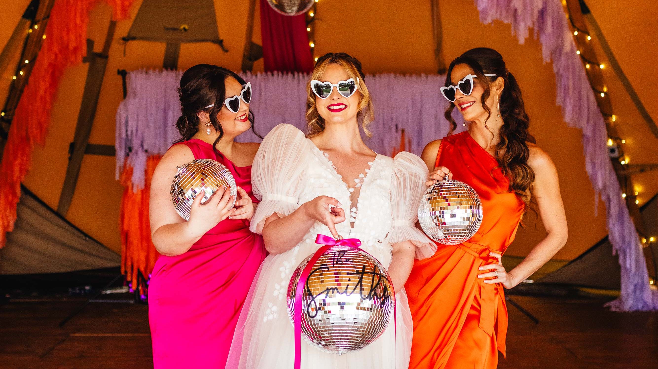 Disco style wedding styling with bright oranges, and pinks, bride holding a disco ball, with bridesmaid either side in neon pink and neon orange dresses, with streamers hanging from the ceiling
