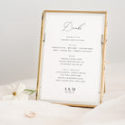 Gold-framed drinks menu on a white surface with a white flower and ring in the background
