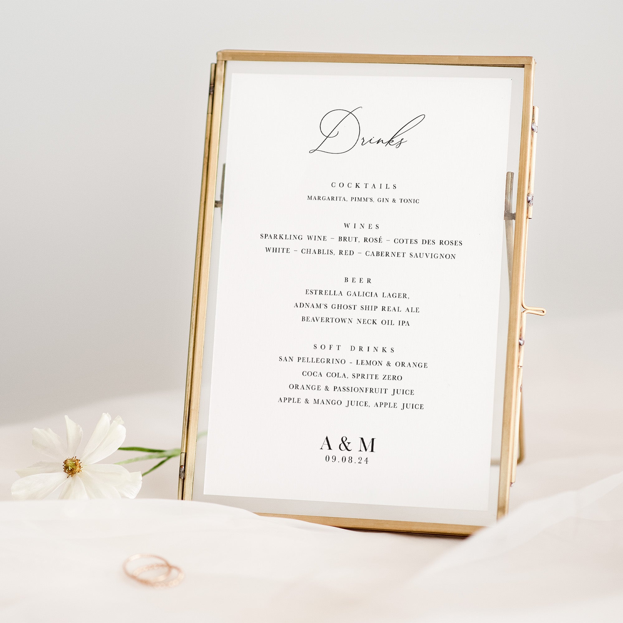 Gold-framed drinks menu on a white surface with a white flower and ring in the background