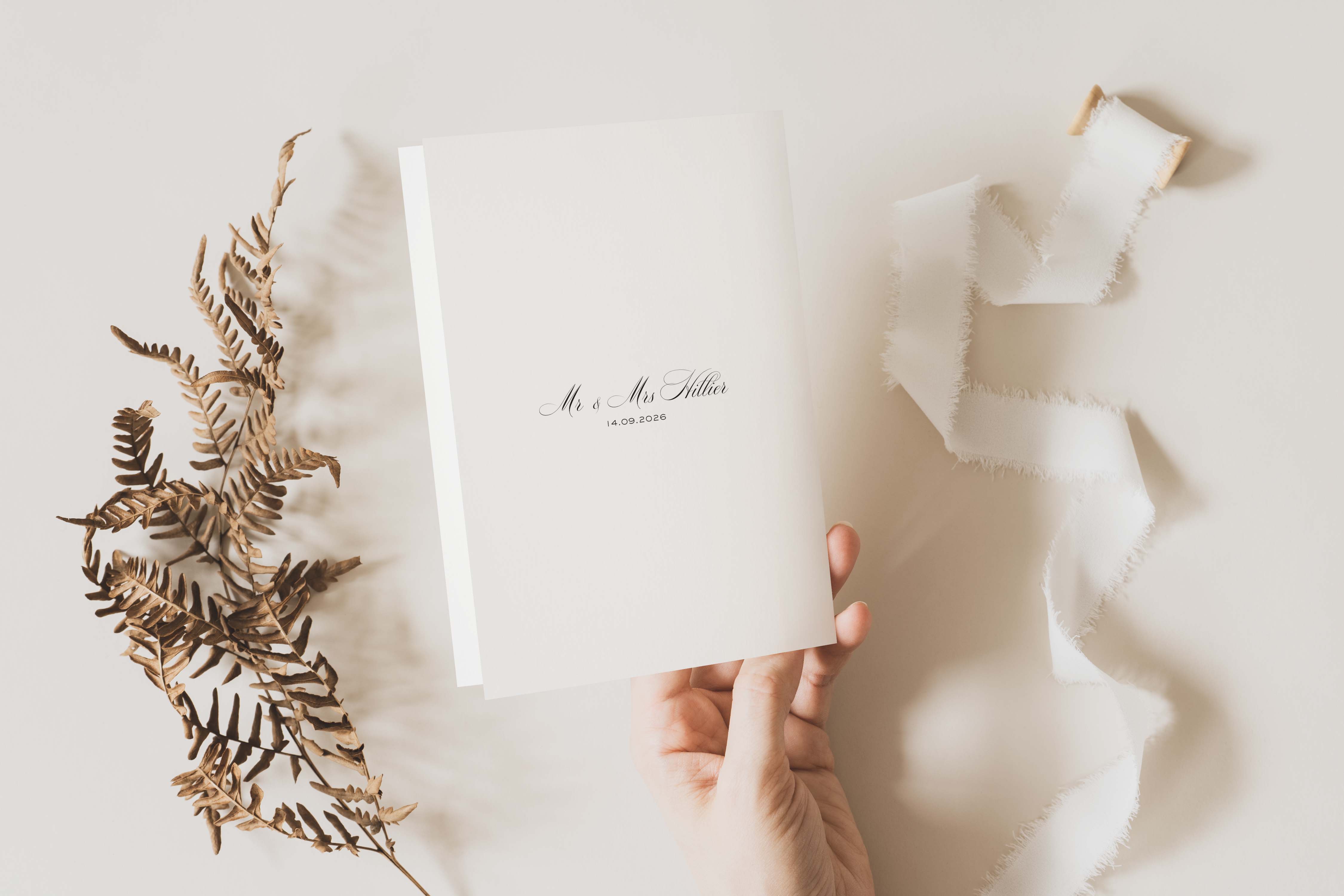 Hand holding a white card with 'Mr & Mrs' text, surrounded by dried plants and fabric on a light background