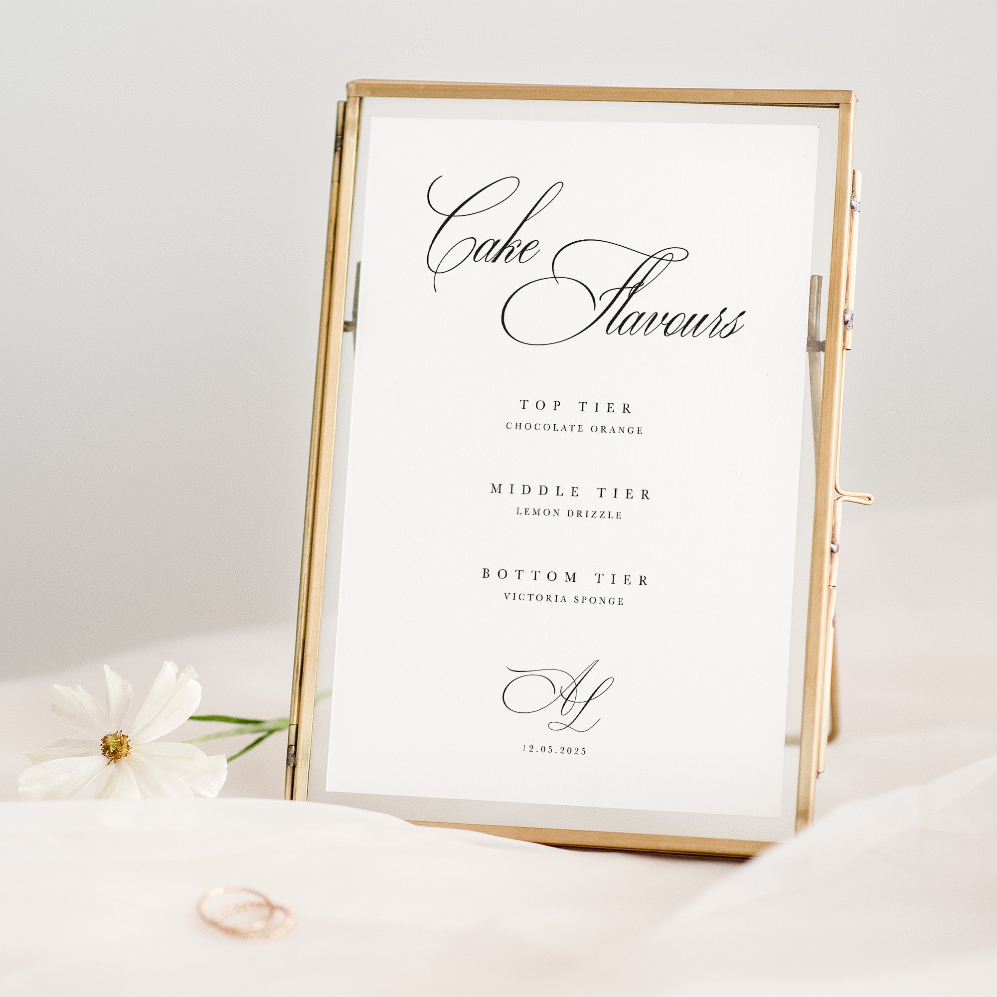 Gold-framed wedding cake flavour sign on a white surface with a white flower and ring in the background.