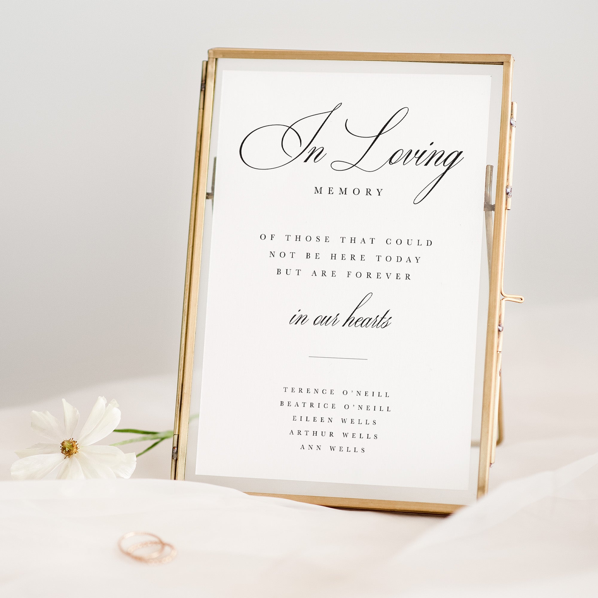 In Loving Memory sign in a gold frame on a white surface with flowers and a ring.