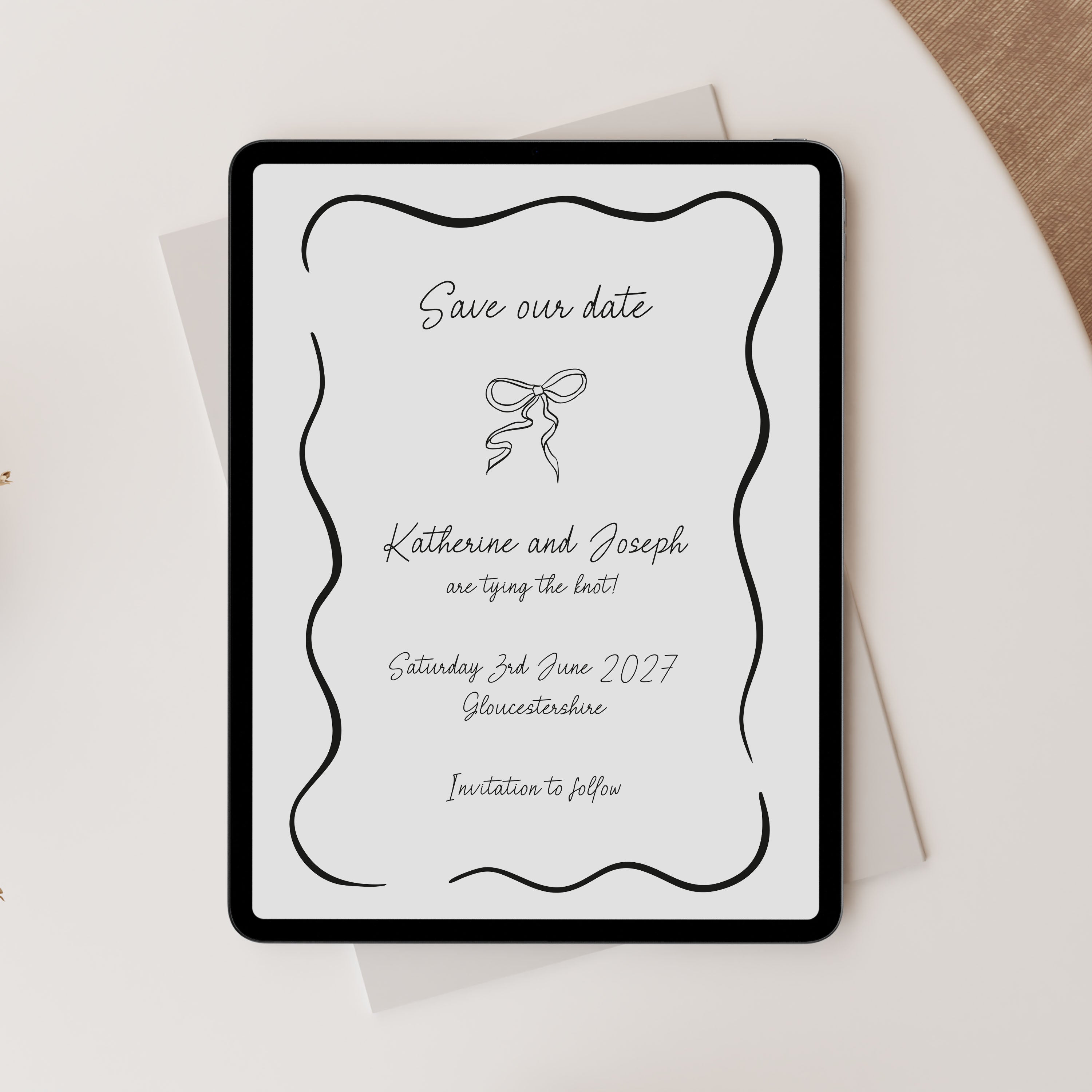 A digital 'Save our date' with elegant black typography and a bow illustration on a white background, styled on top of a light envelope over a cream surface with a beige fabric corner visible.