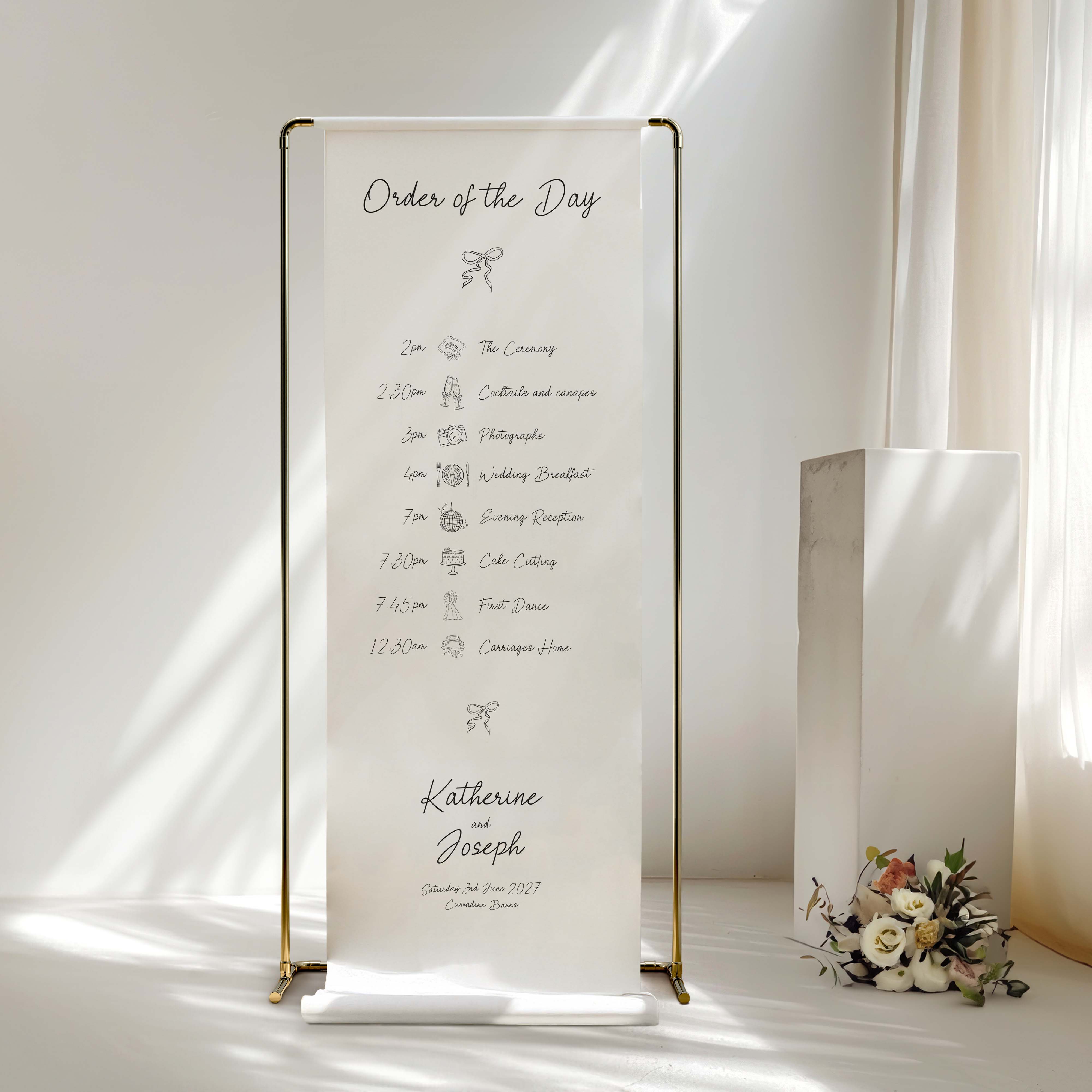 A vertical white fabric wedding order-of-the-day sign with gold metal frame stands on a white floor in a softly lit room. The sign displays a printed timeline from the wedding ceremony to dinner, with the names Katherine and Joseph and the date printed at the bottom.