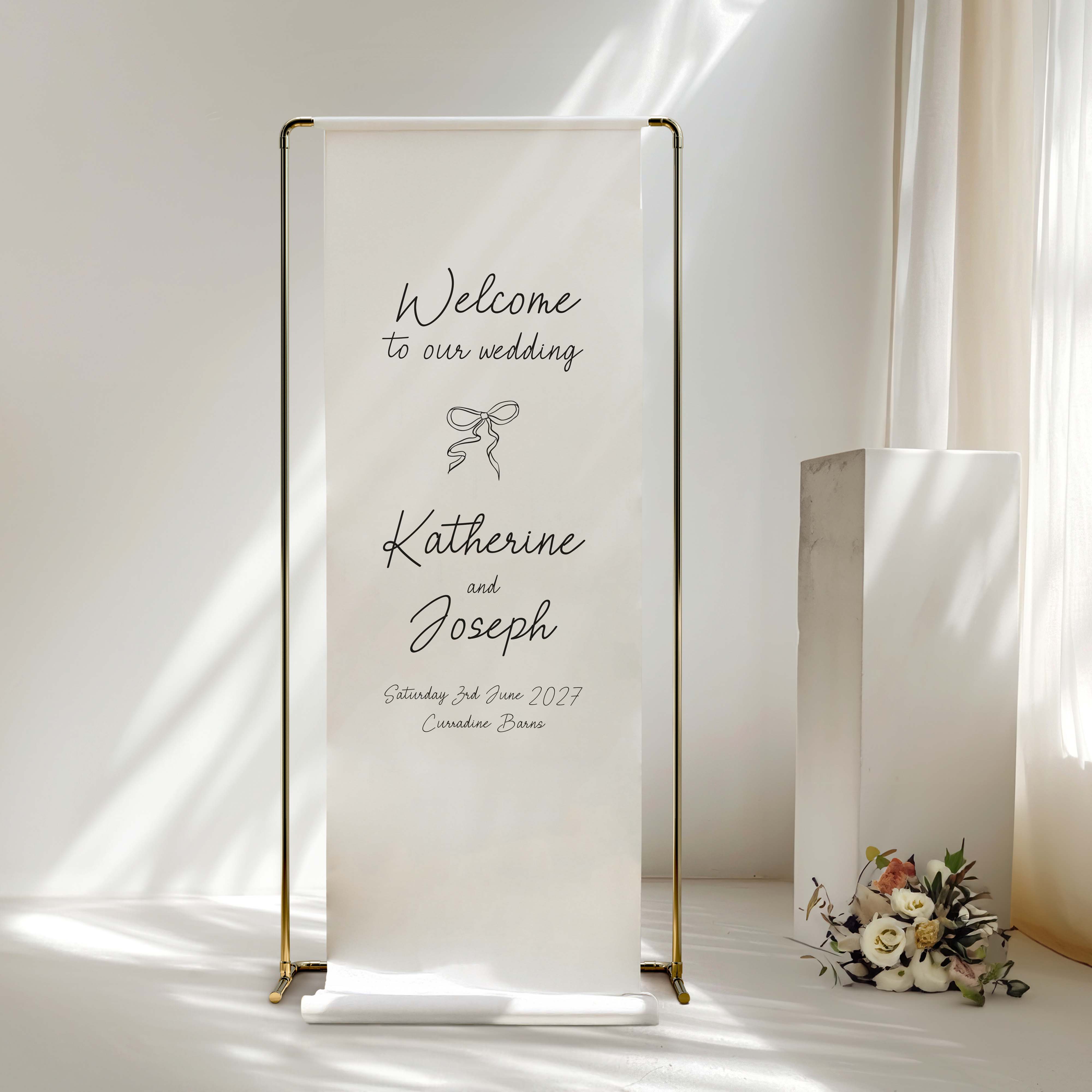 A vertical white fabric wedding welcome sign on a slim gold metal stand, displayed in a bright, sunlit room next to a white pedestal and a small floral arrangement.