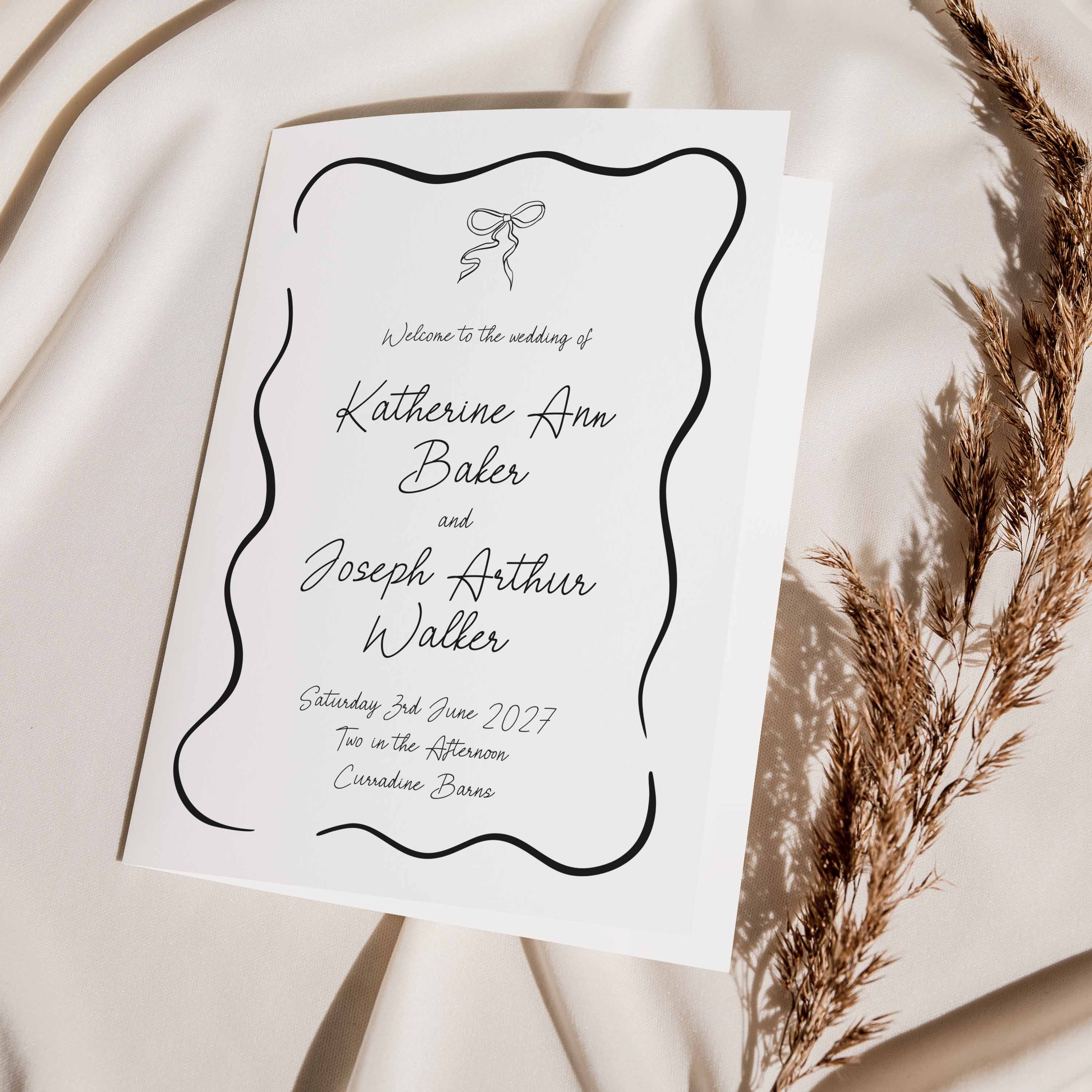 A white wedding order of service booklet with elegant black script text and a decorative wavy border, styled on draped cream fabric with dried pampas grass.