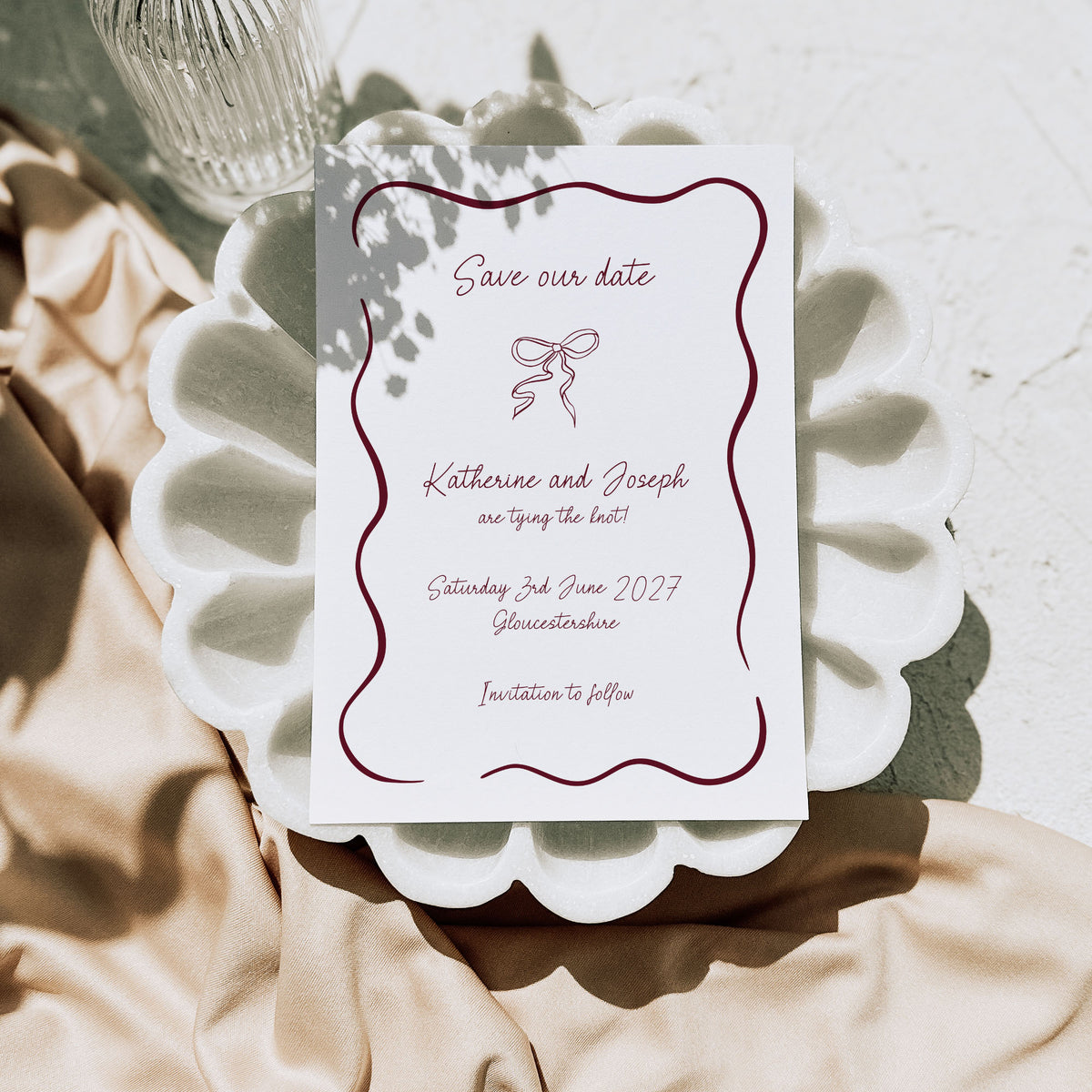 A white save the date card with a whimsical design shown printed in burgundy, sat on top of a stylised white plate, alongside neutral fabric and a glass bottle.