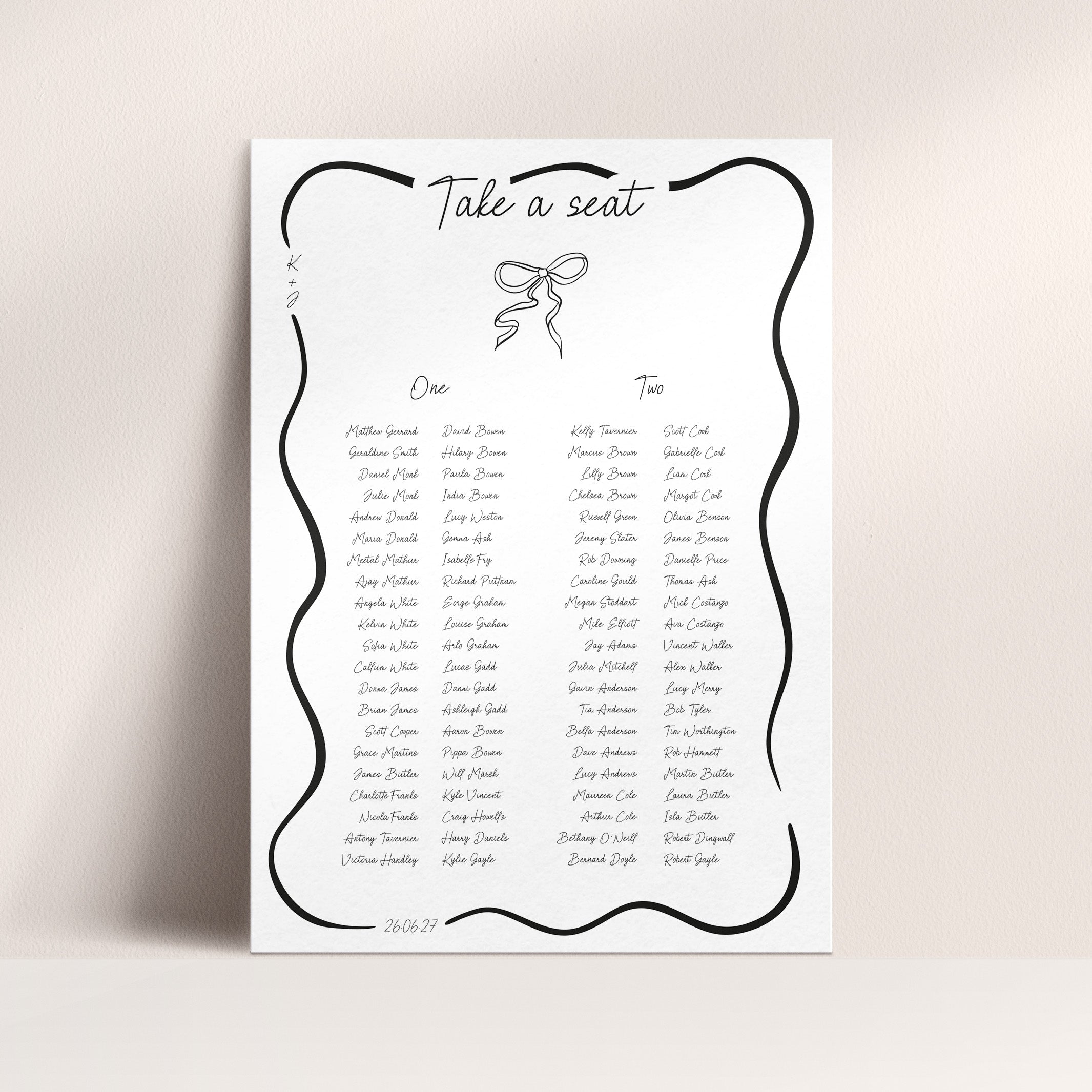 A white wedding seating chart print with a black decorative border, the heading 'Take a seat,' and printed names arranged in columns under labeled sections 'One' and 'Two,' shown against a light beige wall.