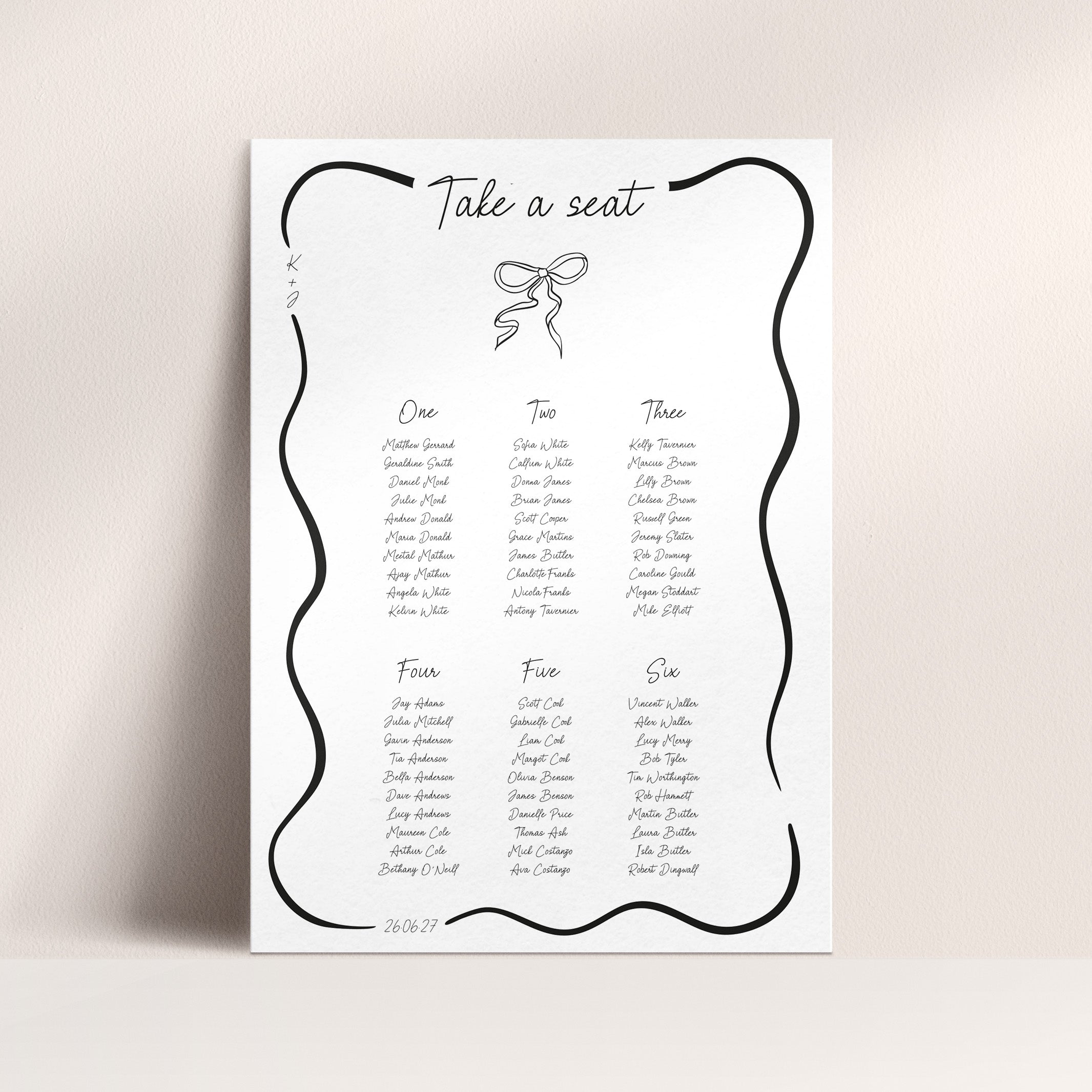 A wedding seating chart print with a decorative black wavy border, the heading 'Take a seat,' and tables for One, Two, Three, Four, Five, and Six printed in black text on a white sheet, shown against a light beige wall.