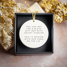 Round white ornament with text in a black gift box with gold ribbon, surrounded by decorative elements.