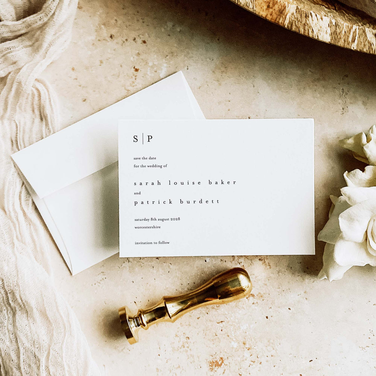 A landscape monogram save the date card in white above a straight flap white envelope, marble background and alongside white foliage, fabric and a gold wax stamp
