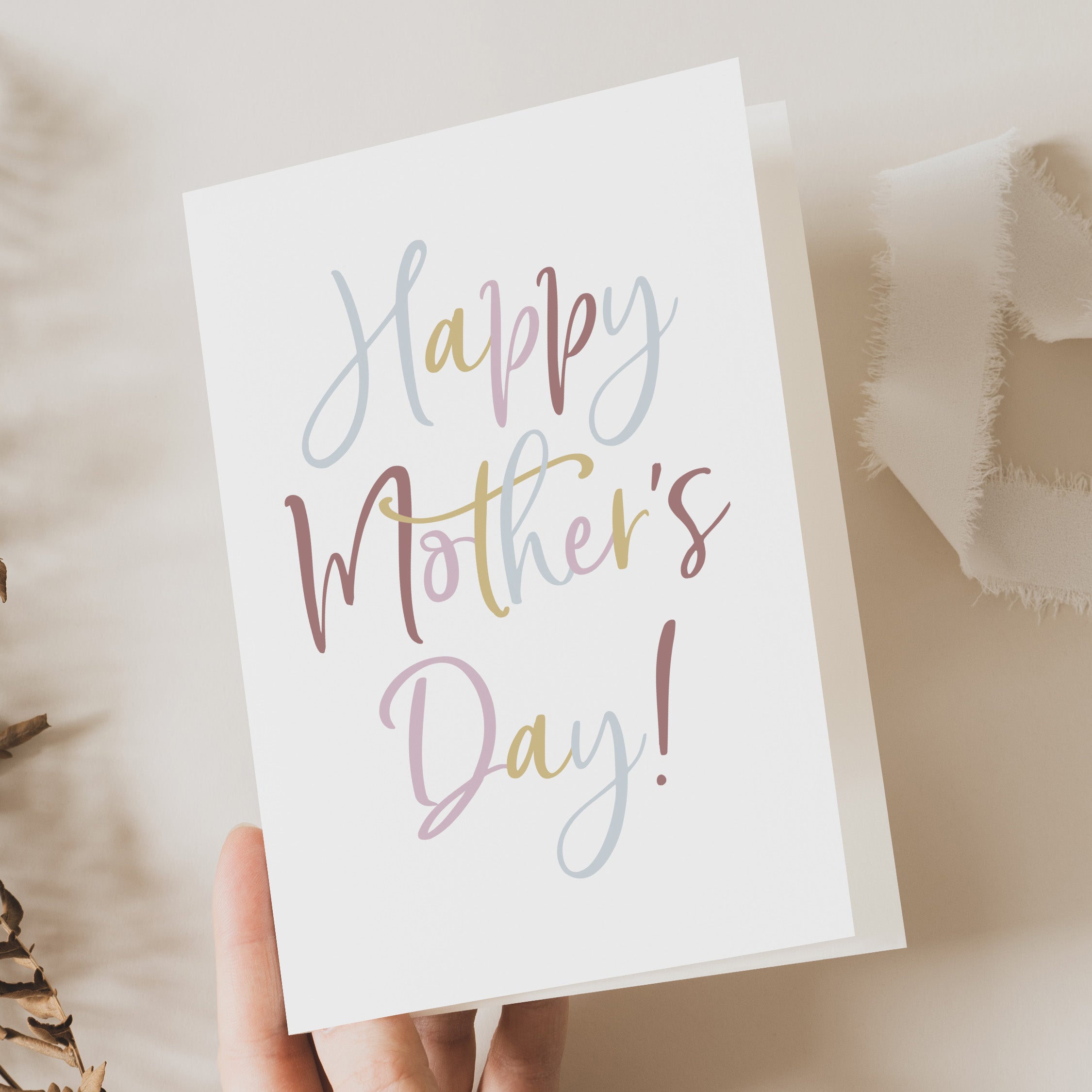 Hand holding a 'Happy Mother's Day!' card on a soft beige background