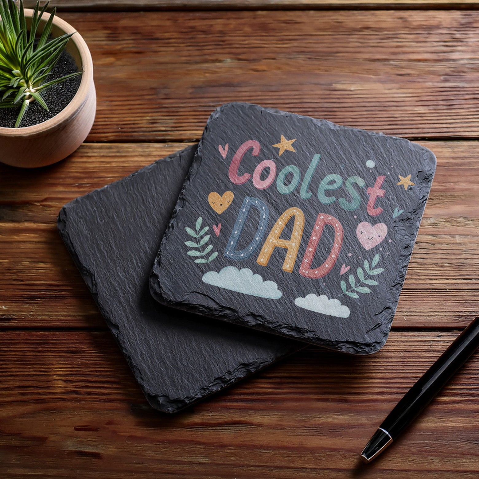Slate coaster with 'Coolest Dad' text on a wooden surface with a pen and plant.