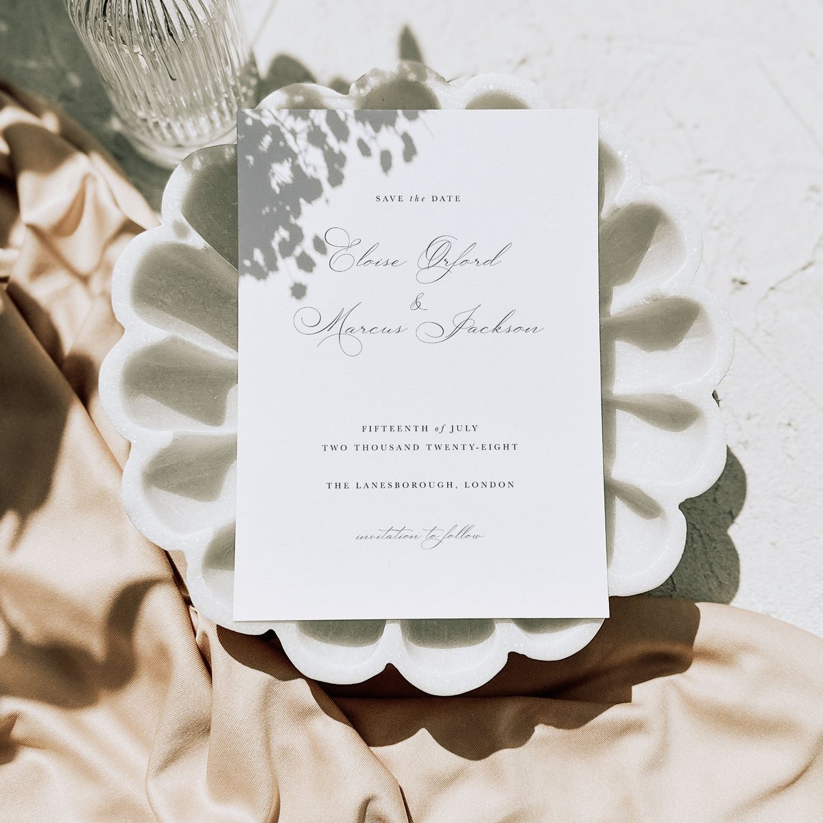 White save the date card with elegant handwritten text, displayed on a white seashell style plate above neutral coloured fabric and alongside a glass bottle
