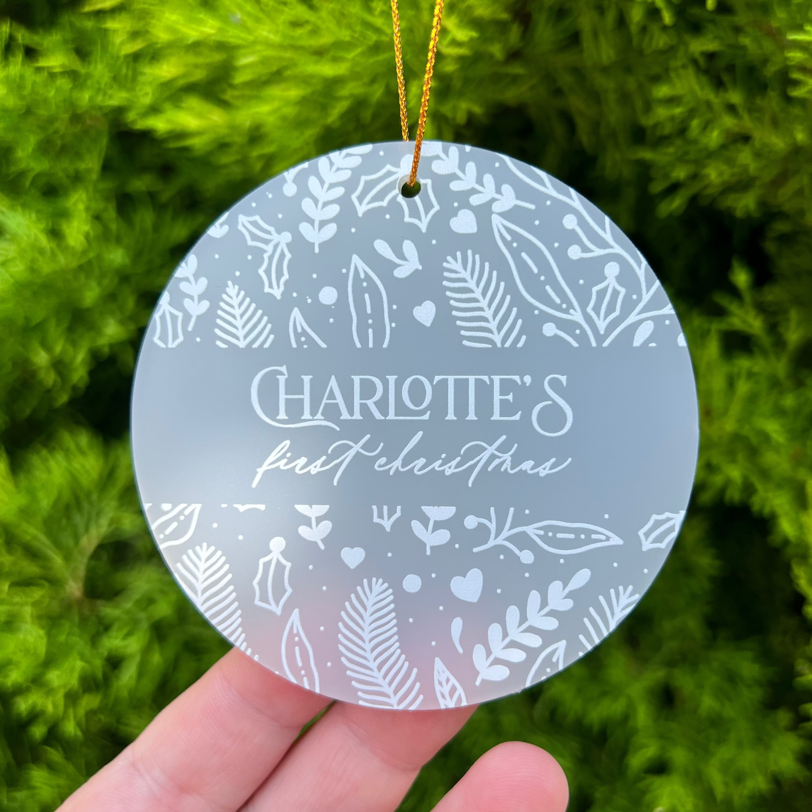 A round frosted glass Christmas ornament with white botanical illustrations and the personalized text 'Charlotte's first christmas', held in a hand against a background of green pine branches.