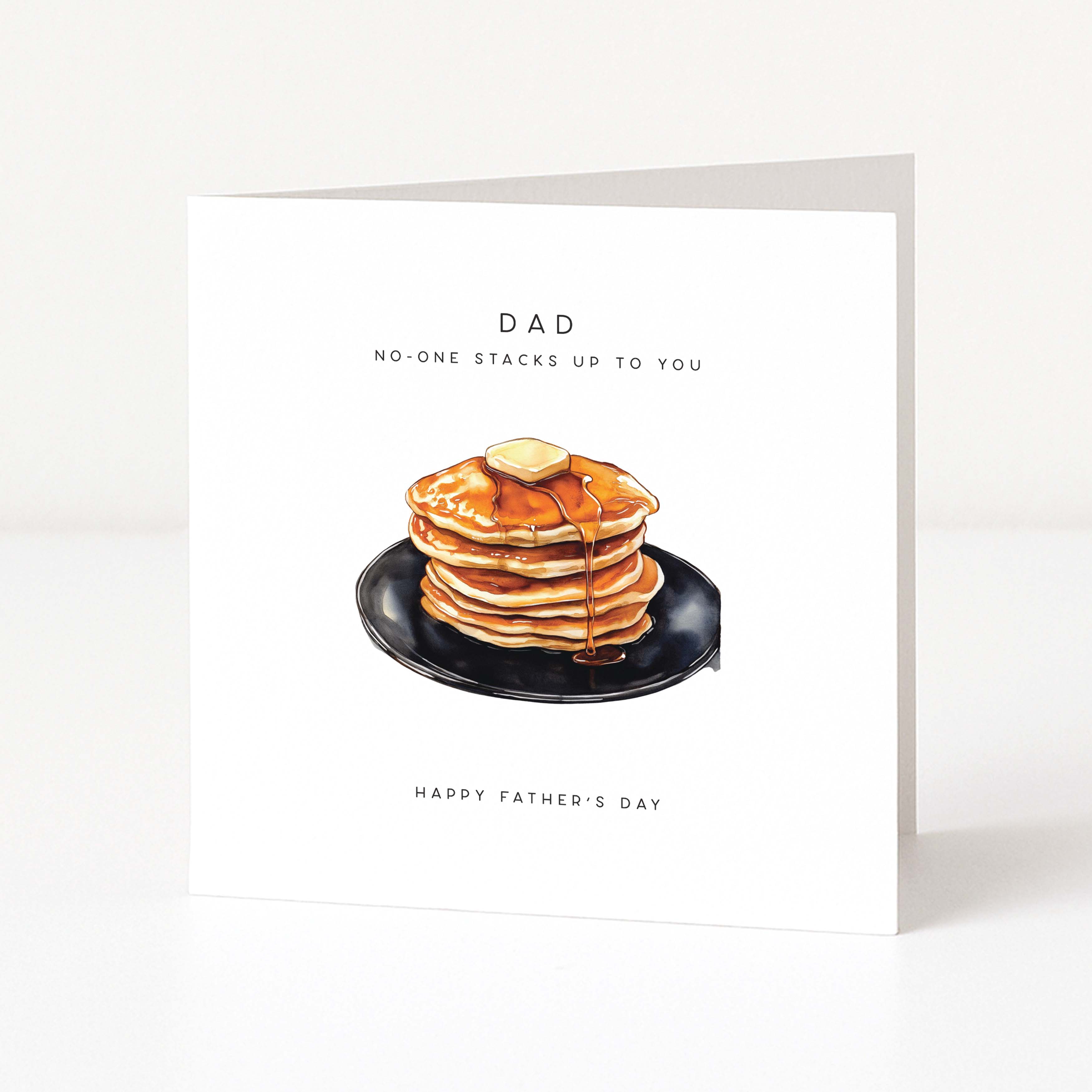 Greeting card with a stack of pancakes and syrup on a black plate, text 'DAD NO-ONE STACKS UP TO YOU' and 'Happy Father's Day' on a white background.