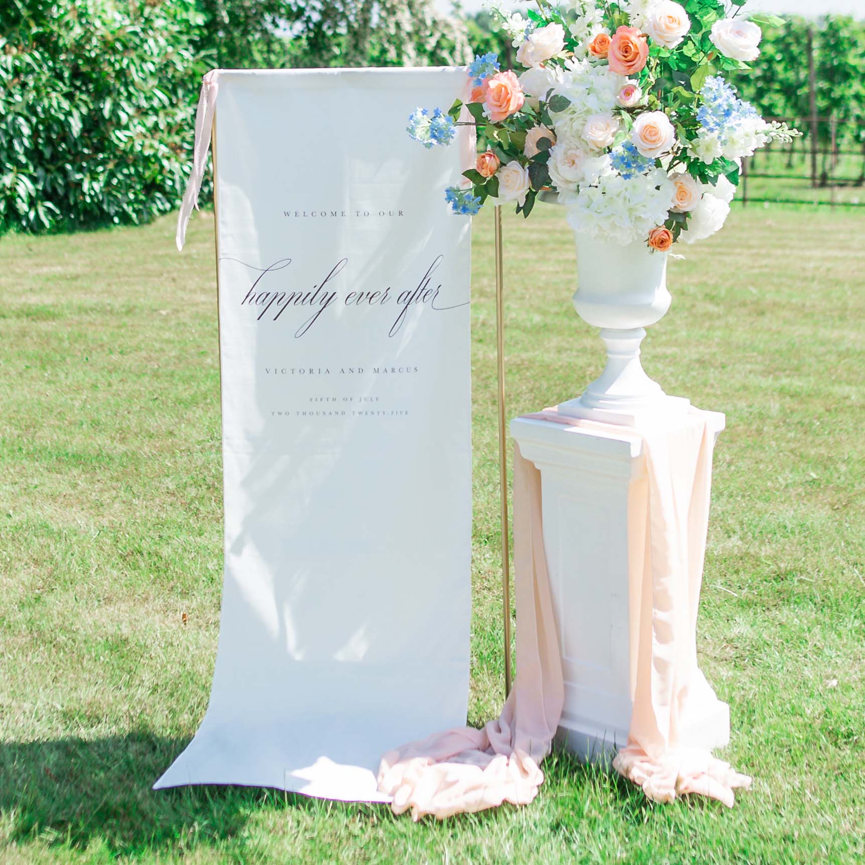 Cotton Linen Wedding Welcome Sign Hanging by a Stand Accompanied by Florals in an Urn