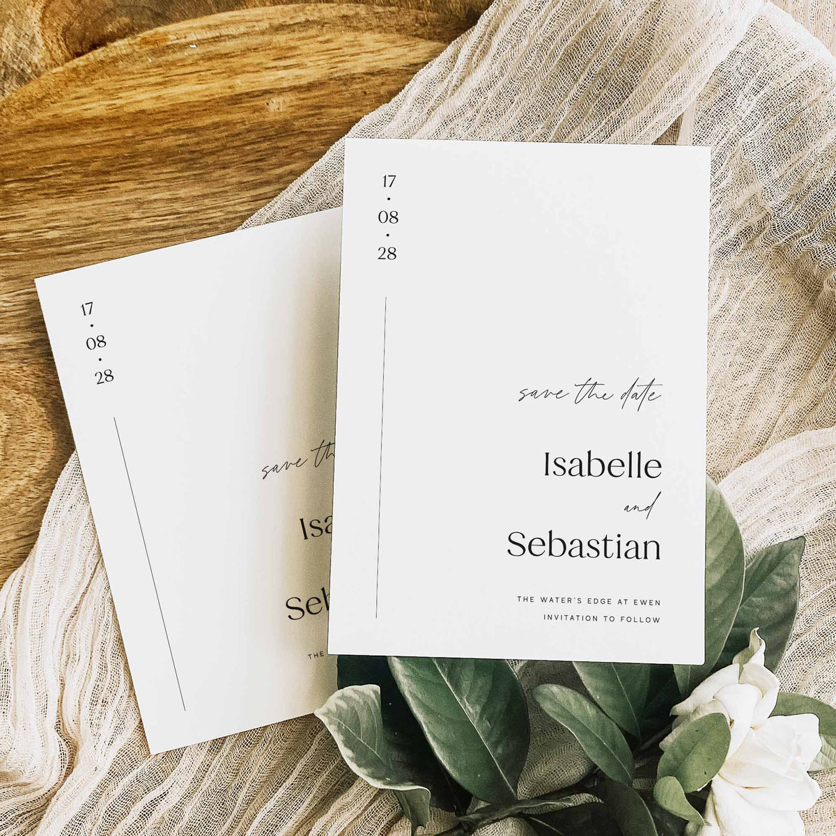 Two white save the date cards for a wedding displayed on sheer white fabric and on a wooden display board, alongside green and white foliage.