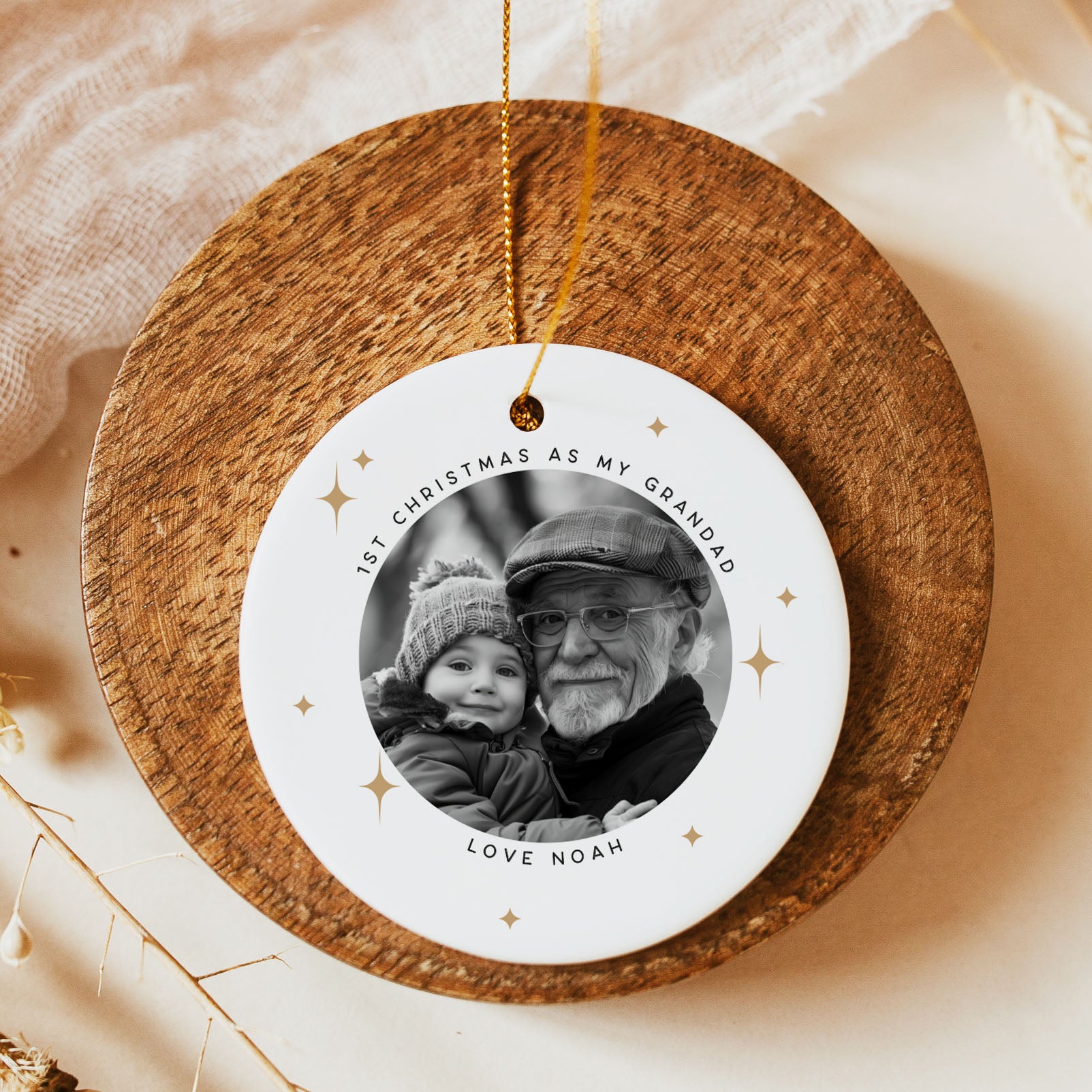 A round white personalized Christmas ornament with a printed black-and-white photo of an older man and child, hanging from a gold string over a rustic wooden board on a warm beige background.