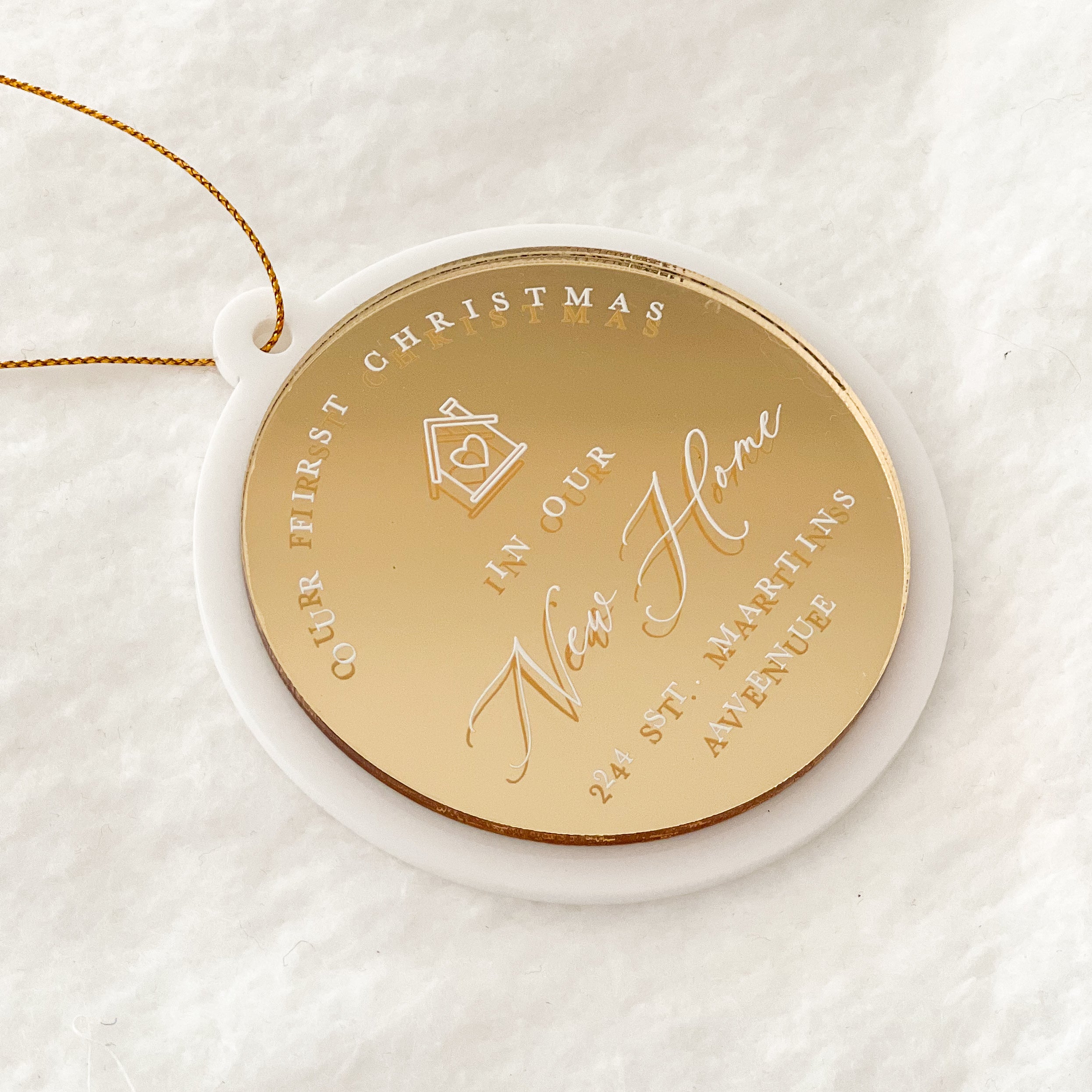 A round gold-toned Christmas ornament with engraved text reading 'Our First Christmas In Our New Home 24 St. Martins Avenue' and a small house icon, displayed on a white circular backing over a soft white fabric background.