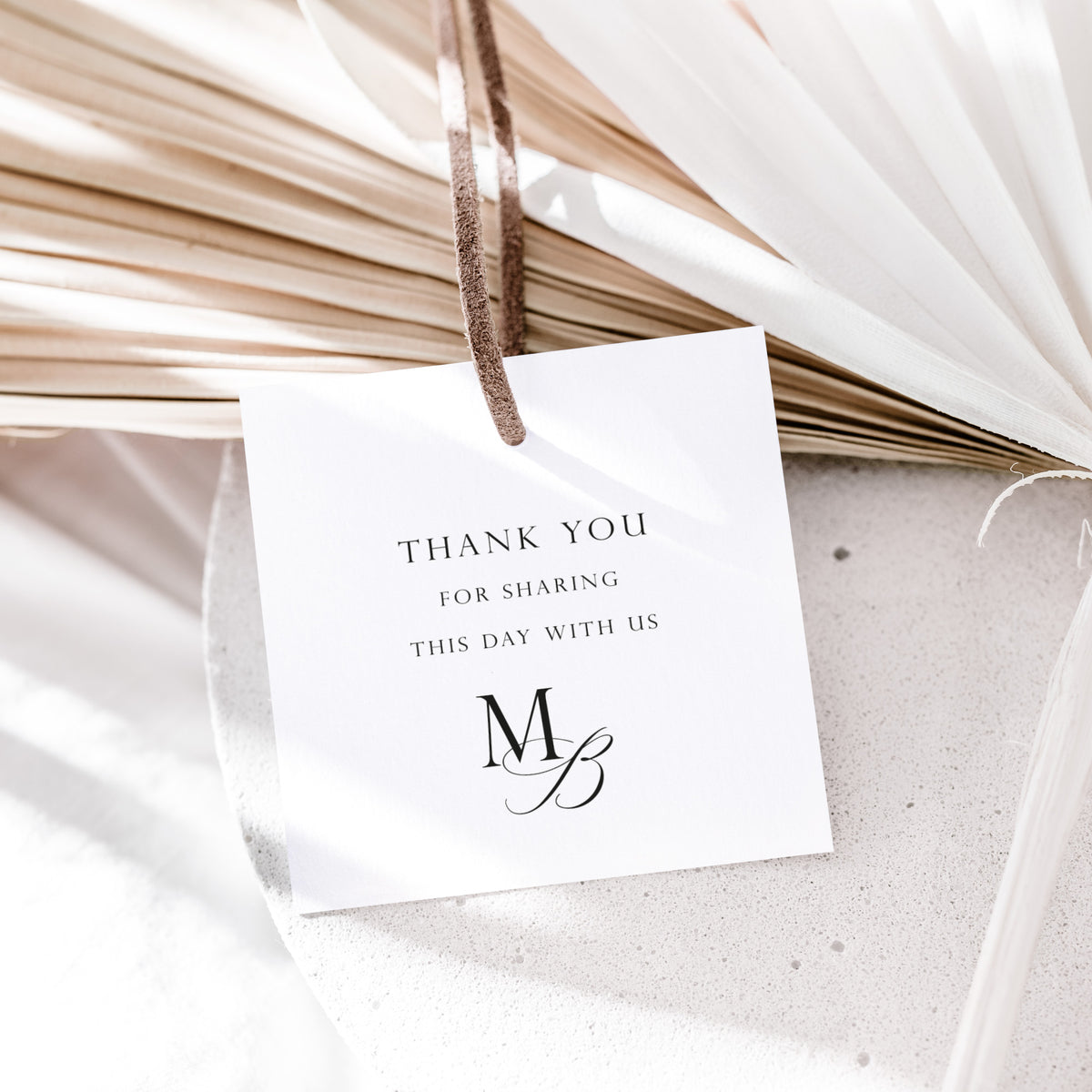 A white square thank-you tag with black text reading 'THANK YOU FOR SHARING THIS DAY WITH US' and a monogram, hanging from a brown string over dried palm leaves and white fabric.