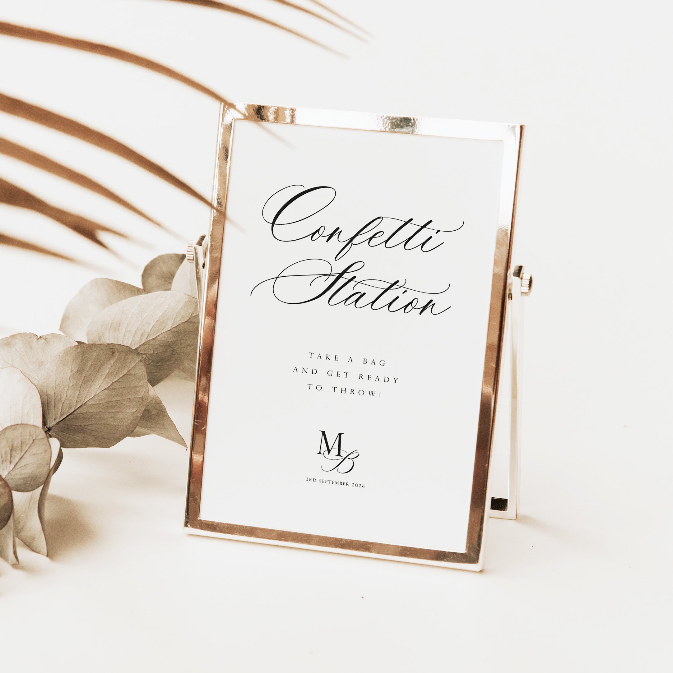 A rectangular freestanding sign with a metallic rose-gold frame and a white insert reading 'Confetti Station' in black script, styled on a light surface with dried leaves and palm fronds.