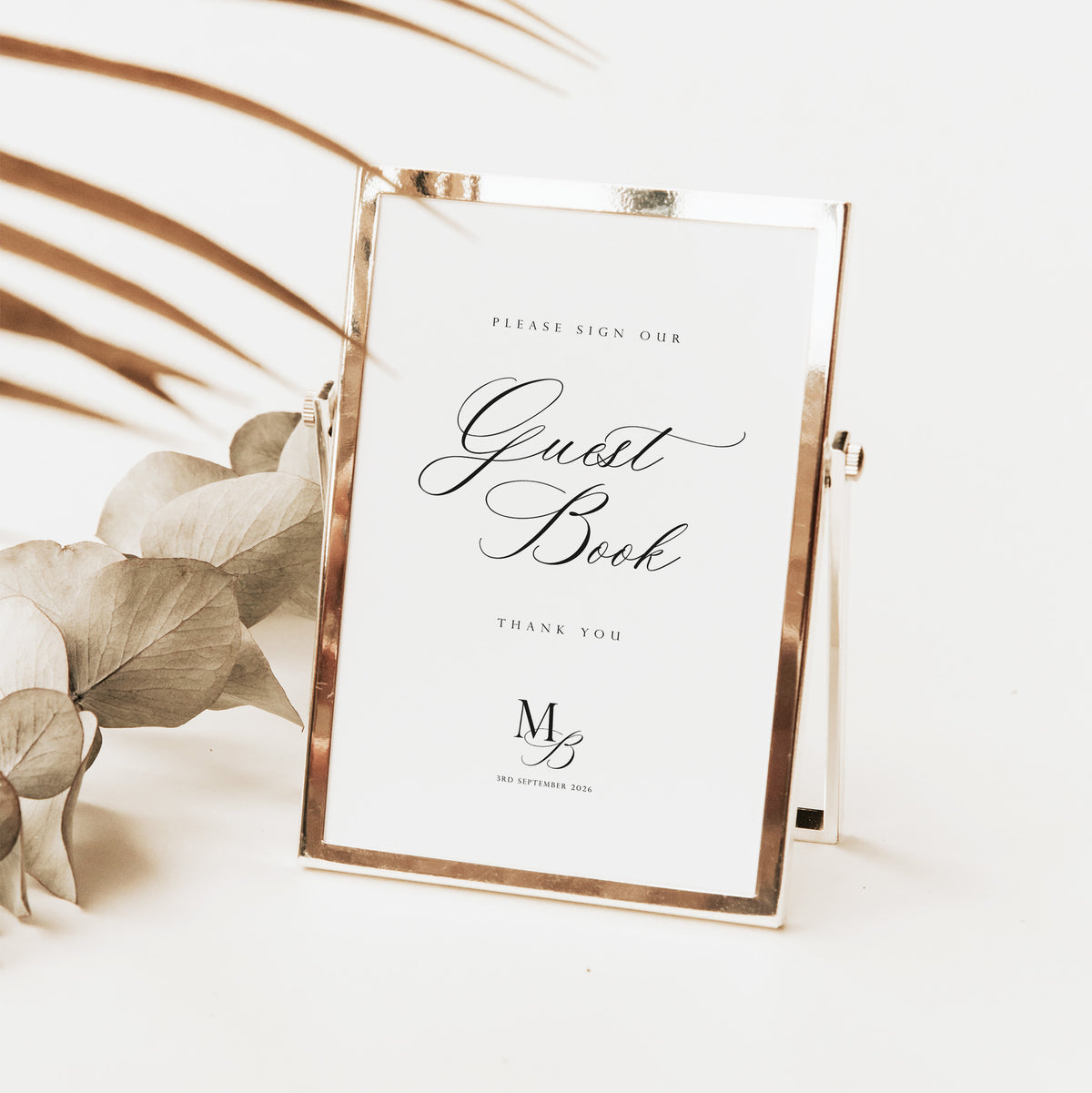 A rose gold metal frame guest book stand with a white insert, styled on a white surface with dried palm leaves and eucalyptus flowers.