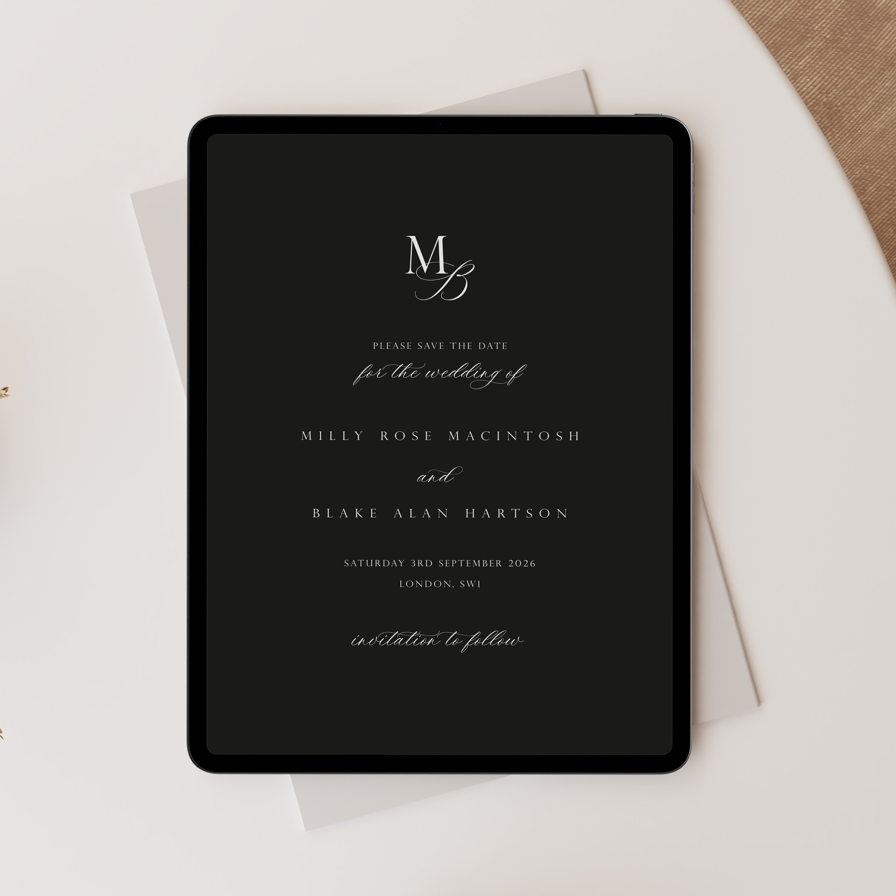 A black tablet with elegant white wedding invitation text and monogram lettering, styled on top of white envelopes on a light neutral surface with a beige fabric corner visible.
