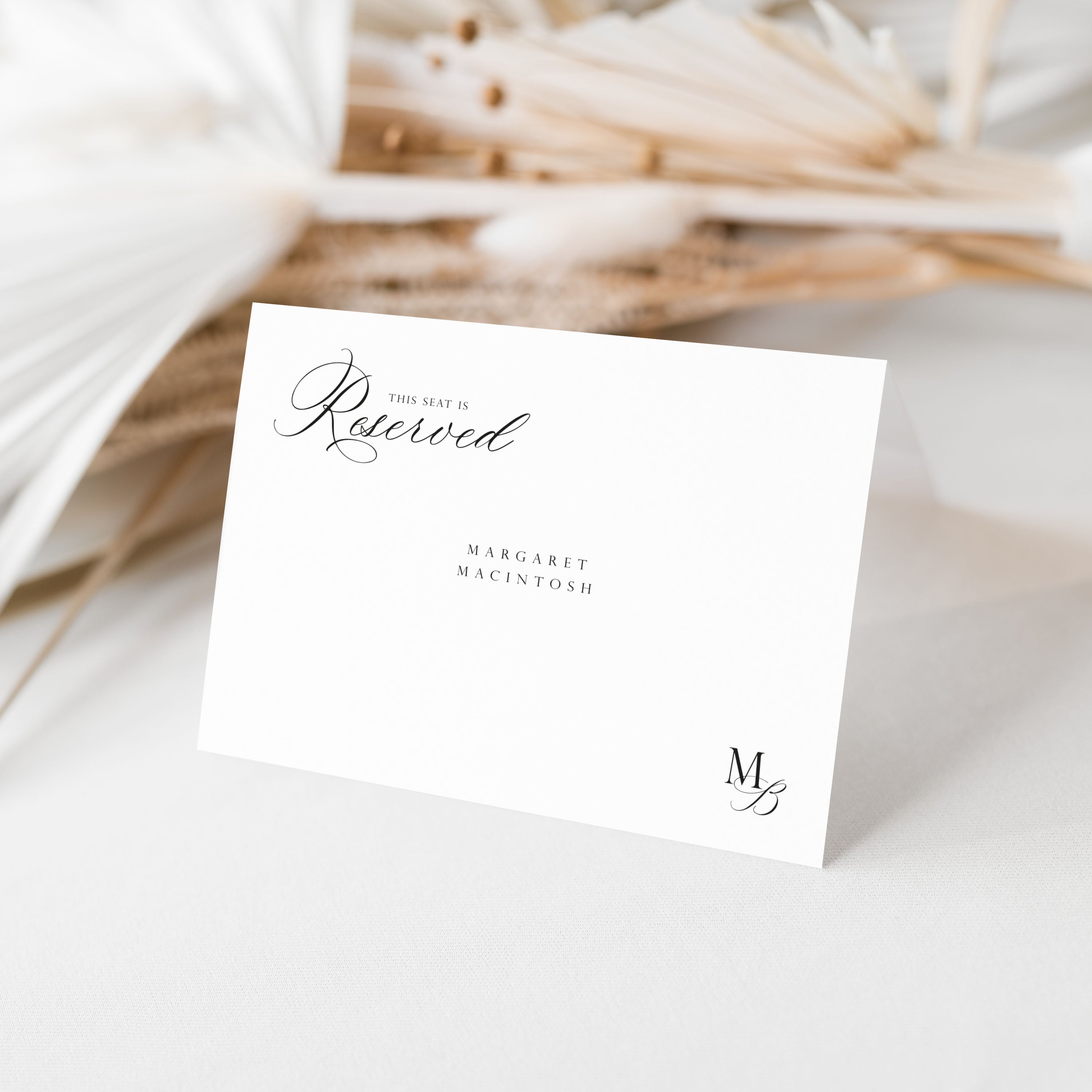 A white reserved seating card with elegant black script text reading 'This seat is reserved' and the names 'Margaret Macintosh,' photographed standing upright on a light surface with soft dried floral decor in the background.
