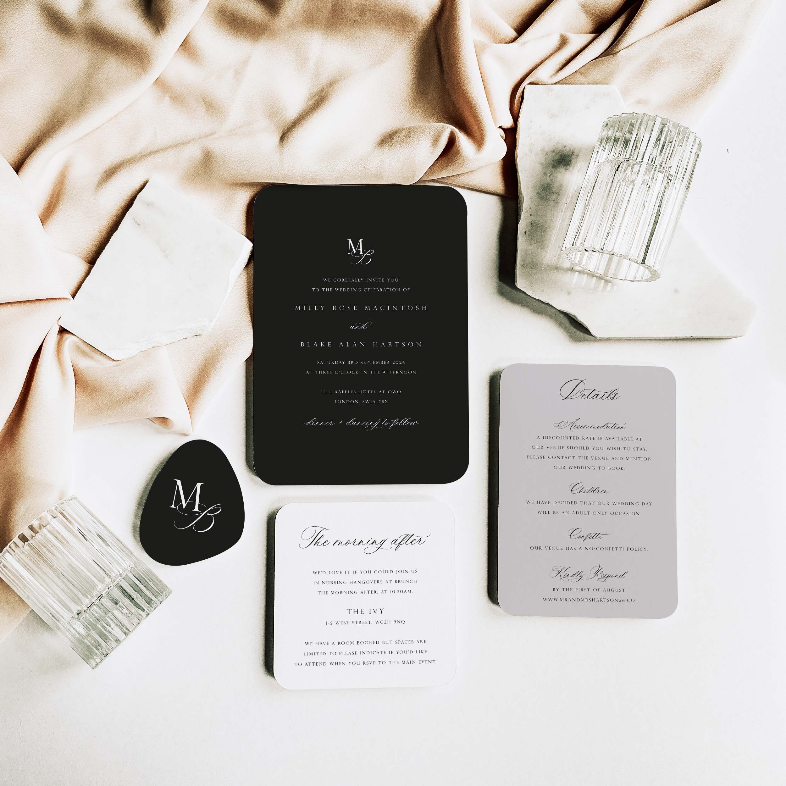 A styled flat-lay of a wedding invitation suite on a white surface with draped blush fabric. The set includes a black invitation card, a white 'The morning after' card, a light grey details card, a black tag with an MB monogram, and clear ribbed glasses.
