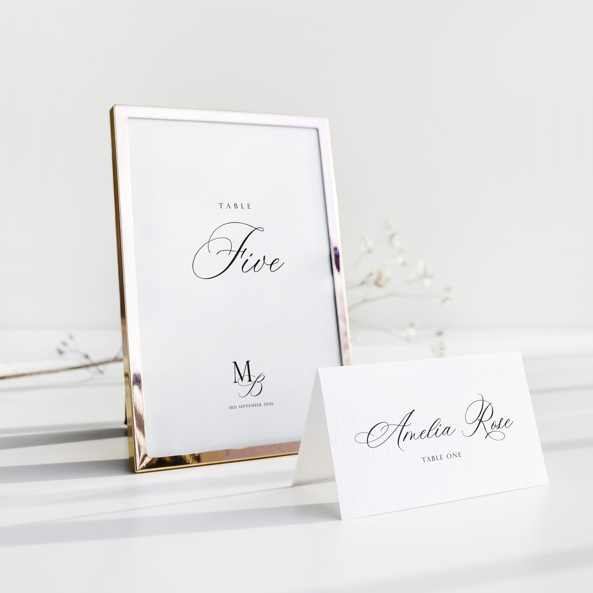 A rose gold metal table number frame standing upright next to a white paper table card on a clean white surface with soft decorative branches in the background.
