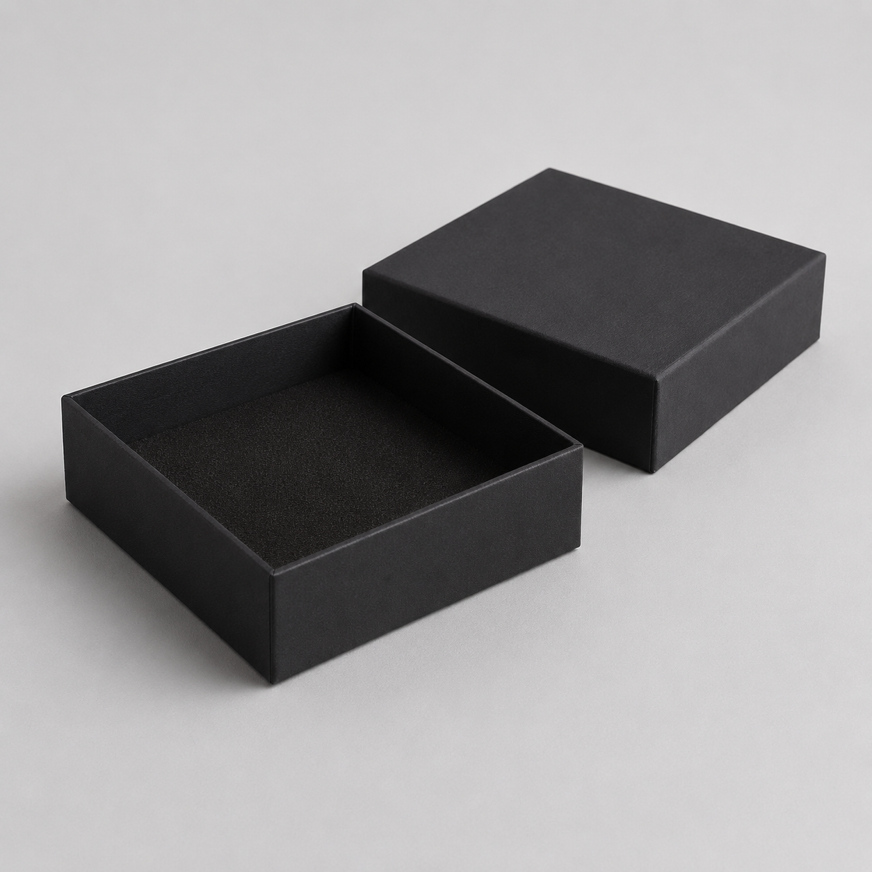 A pair of matte black square gift boxes, one open to show the lined interior and one closed, set against a neutral light gray background.
