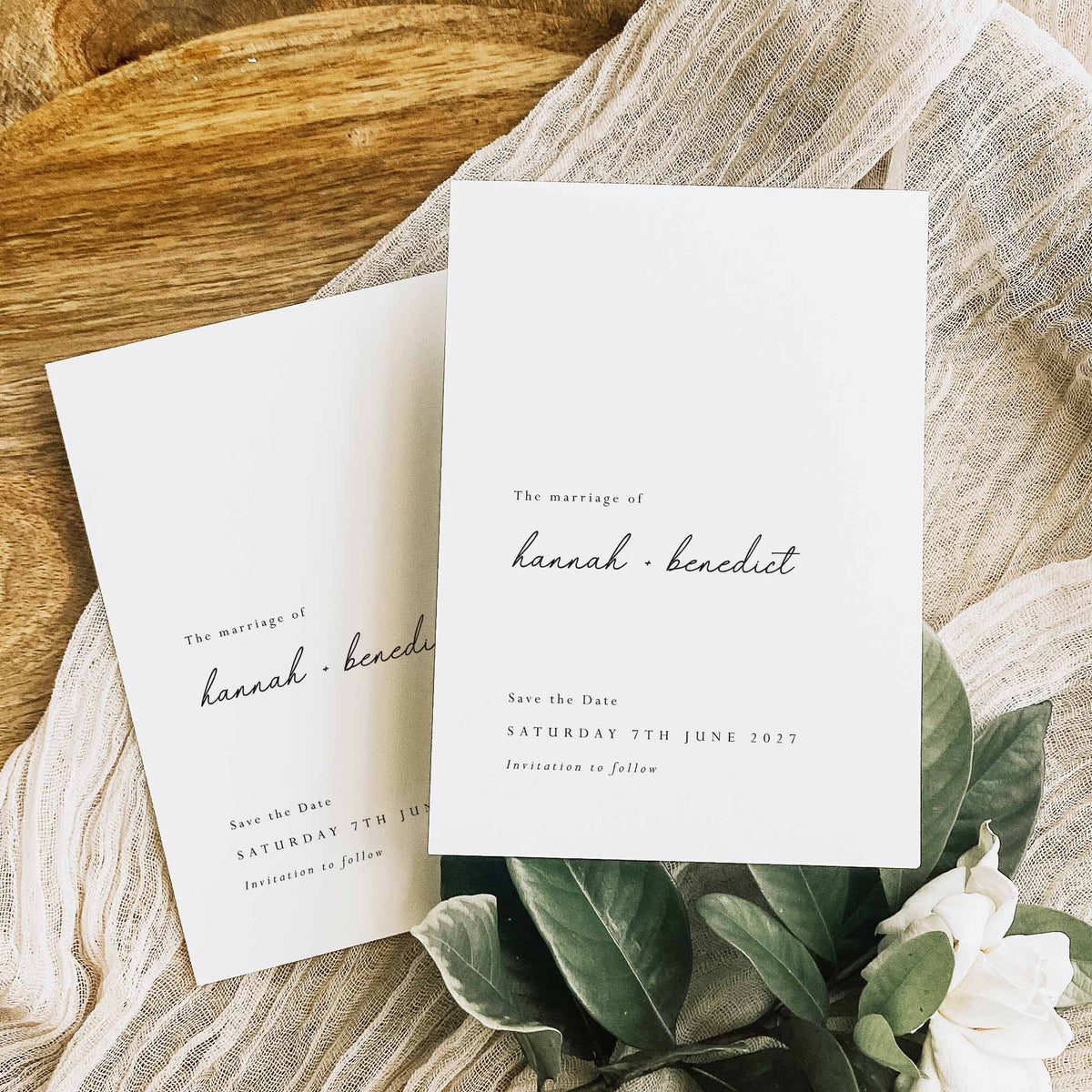 Two white save the date cards displayed on white fabric and a wooden board alongside green foliage.
