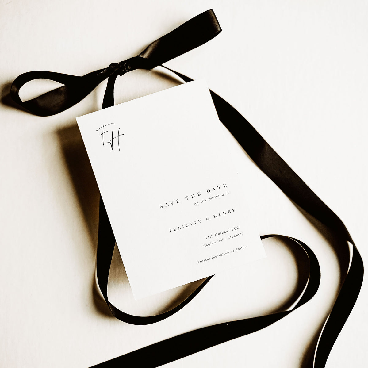 A white save the date card with a monogram and classic text, shown on a white background and alongside a black ribbon tied into a bow.