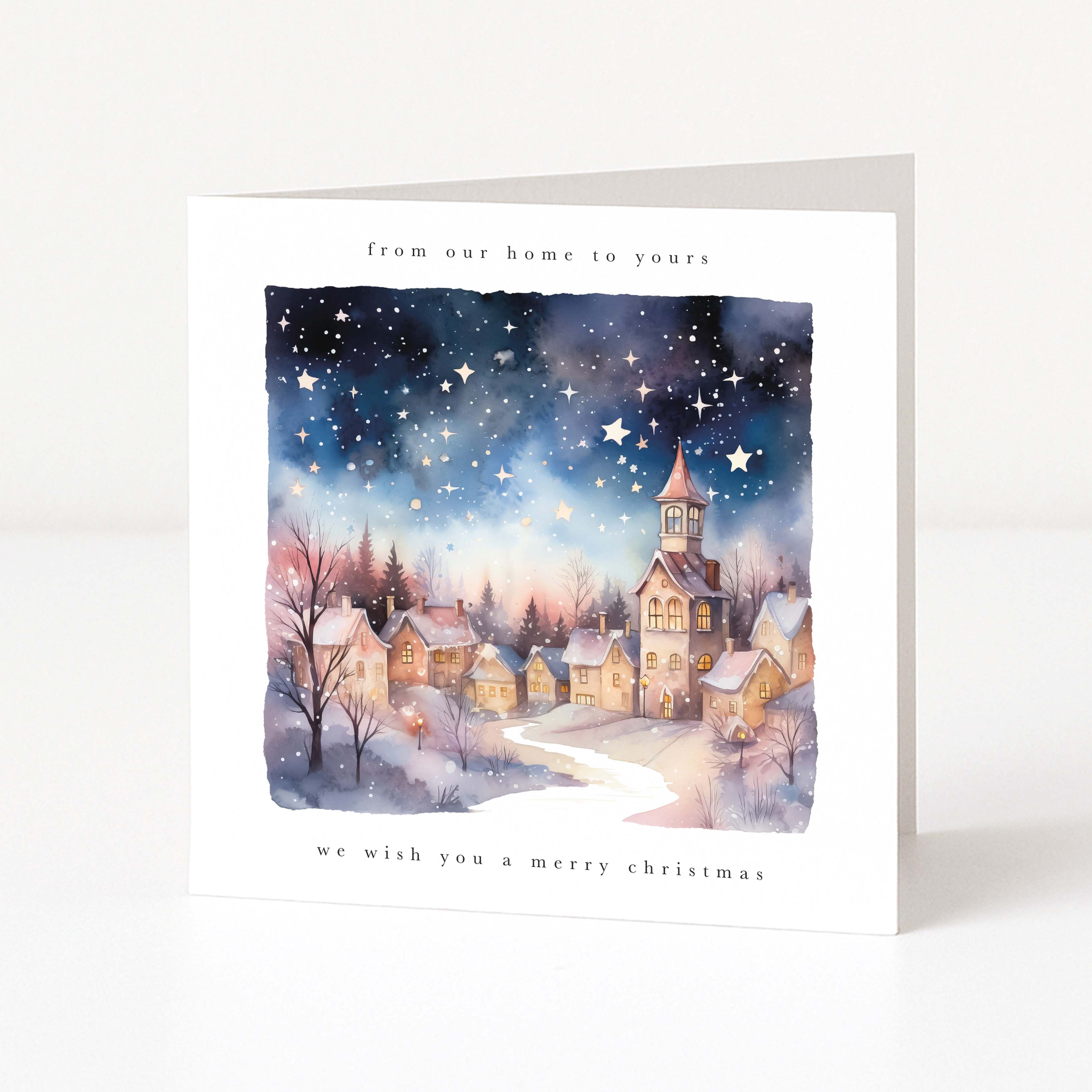 A white Christmas greeting card standing upright on a light gray surface. The front features a watercolor illustration of a snowy village at night under a starry sky, with the text 'from our home to yours' at the top and 'we wish you a merry christmas' at the bottom.