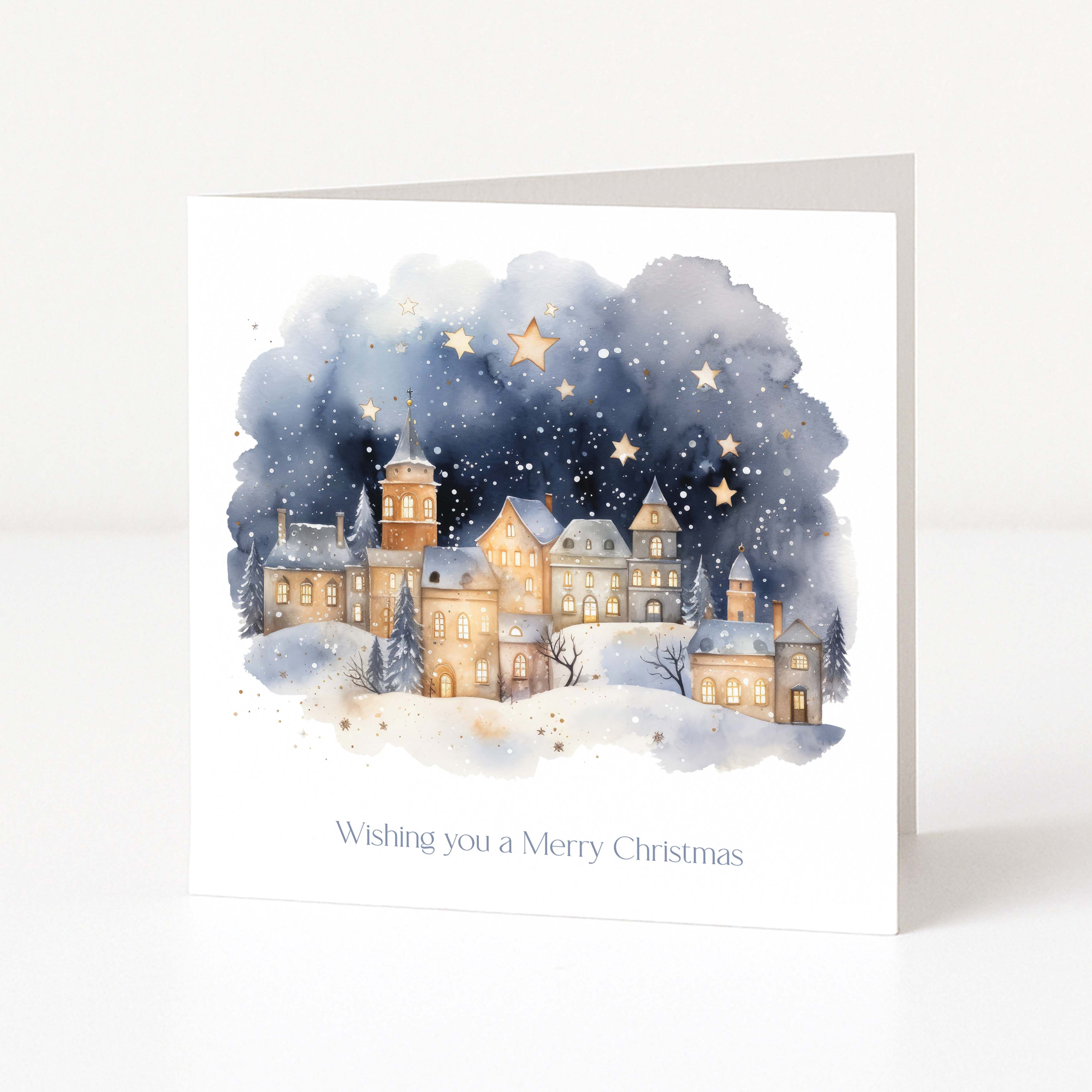 A square white greeting card standing upright on a light surface, printed with a watercolor winter village scene under a starry night sky and the text 'Wishing you a Merry Christmas'.