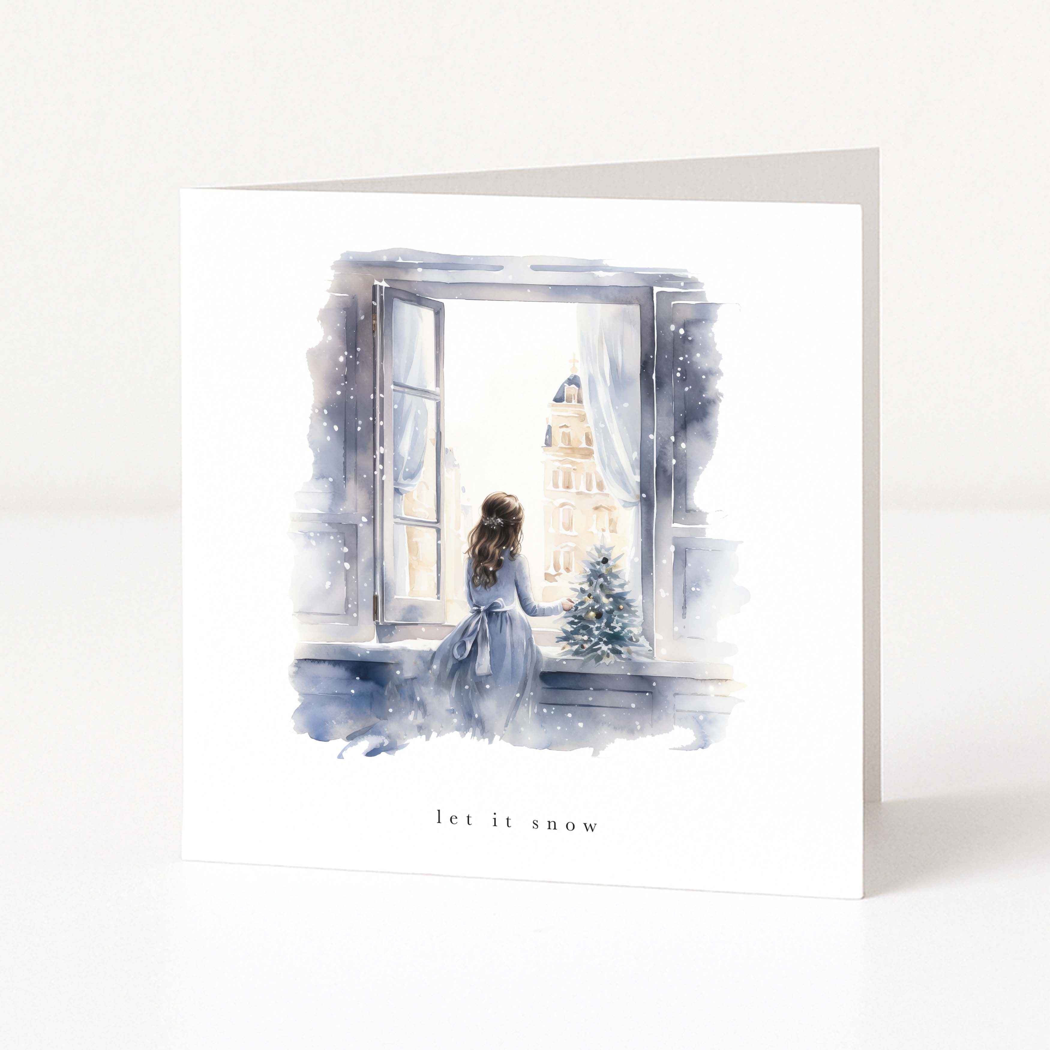 A white greeting card standing upright on a white surface, featuring a watercolor illustration of a woman in a blue dress sitting by an open window and looking out at a snowy city scene. The card is labeled 'let it snow' at the bottom.