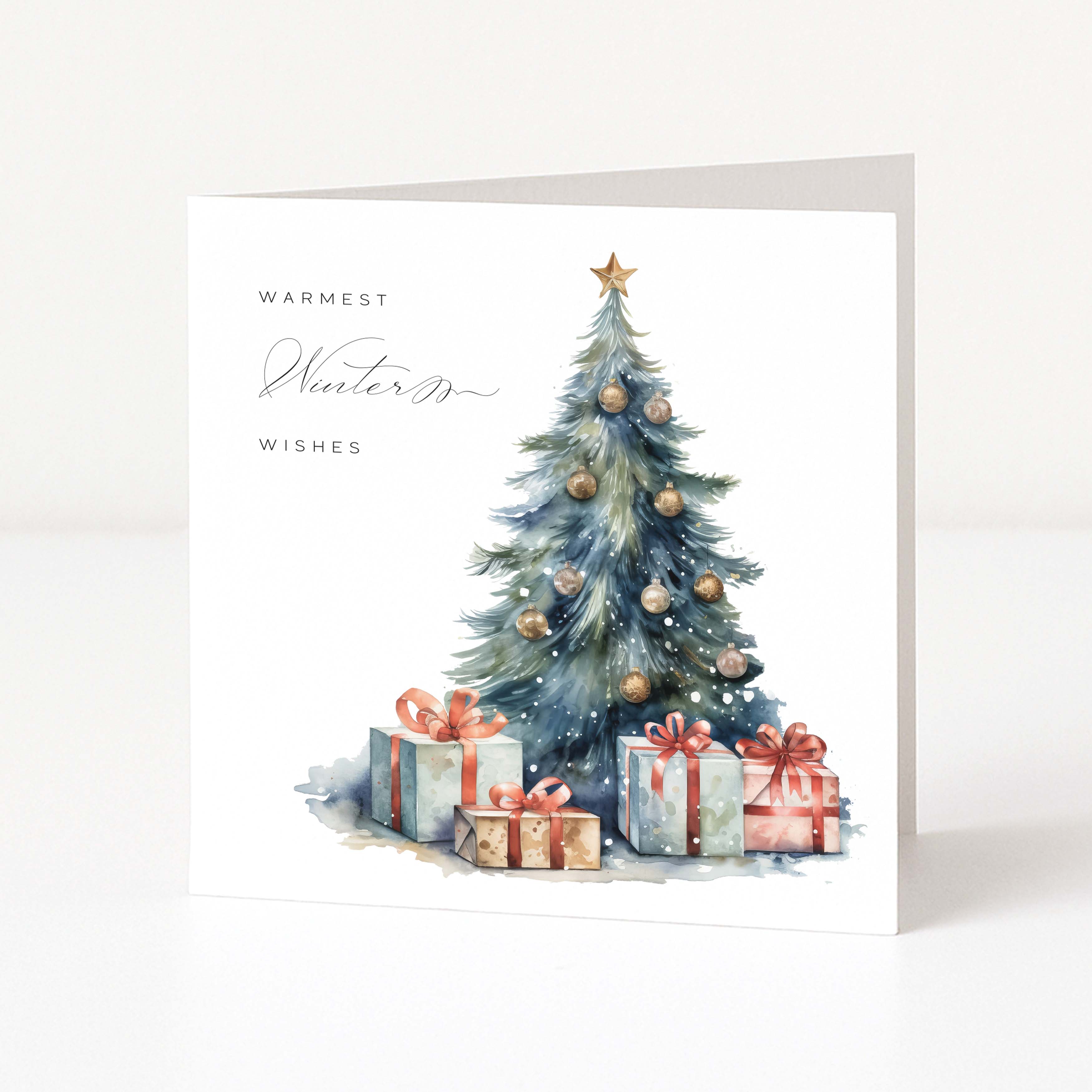 A square white Christmas card standing upright on a light surface, featuring a watercolour Christmas tree with ornaments and wrapped presents beneath, plus the text 'WARMEST Winter Wishes' in black script.