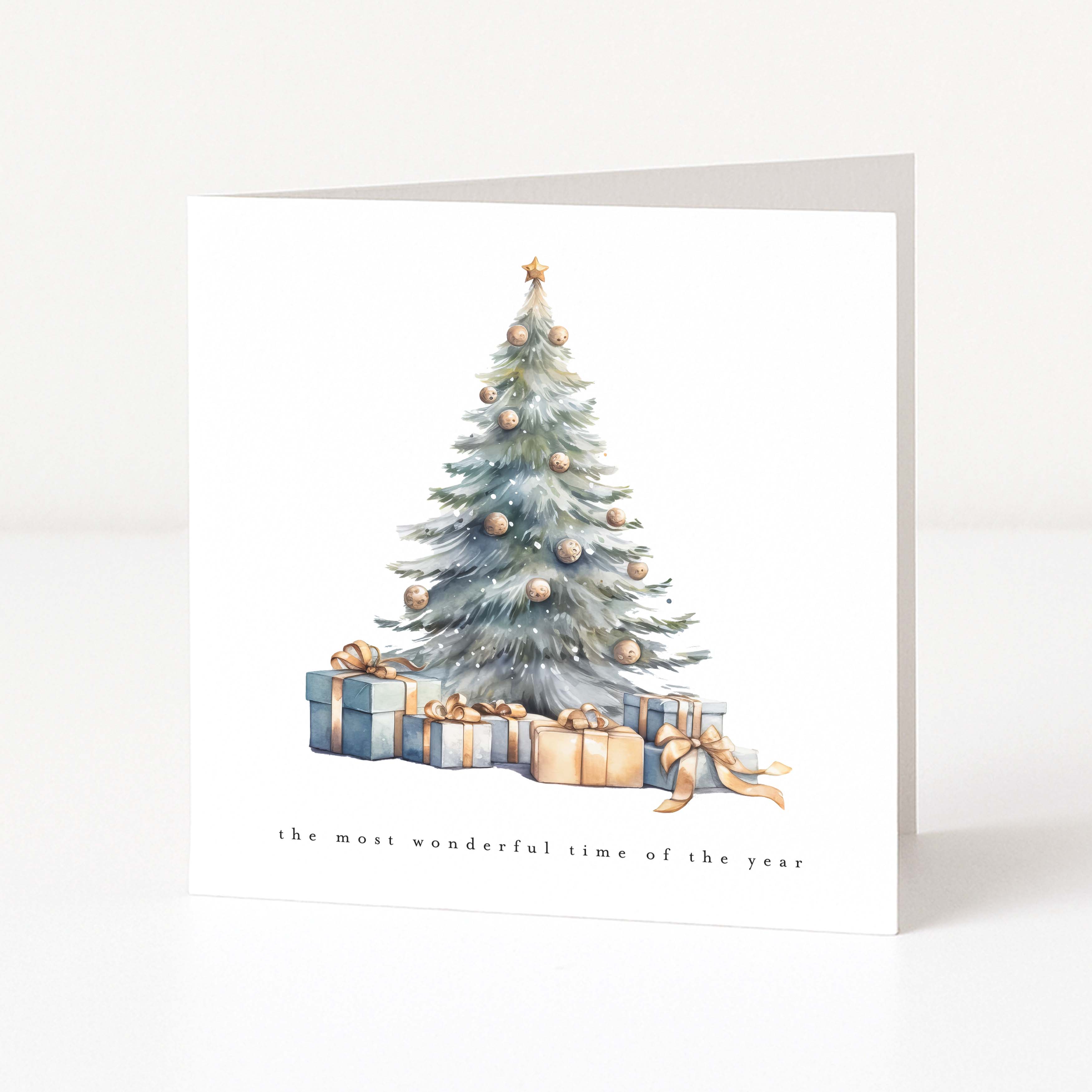 A square white Christmas greeting card standing upright on a light grey surface, featuring a watercolour illustration of a decorated Christmas tree with wrapped presents underneath and the text 'the most wonderful time of the year' near the bottom.