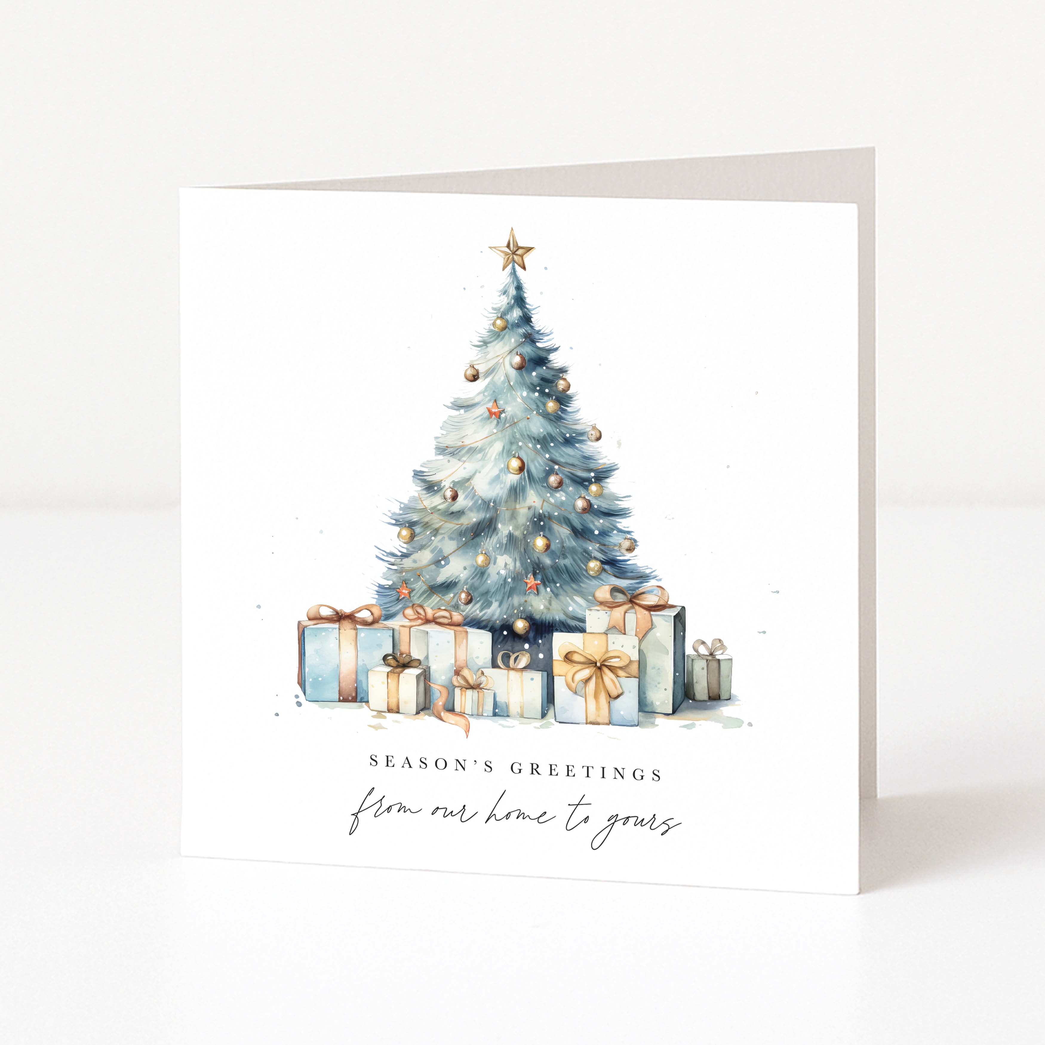A square white Christmas greeting card standing upright on a white surface, featuring a watercolor illustration of a decorated Christmas tree with wrapped presents and the text 'SEASON'S GREETINGS' and 'from our home to yours'.