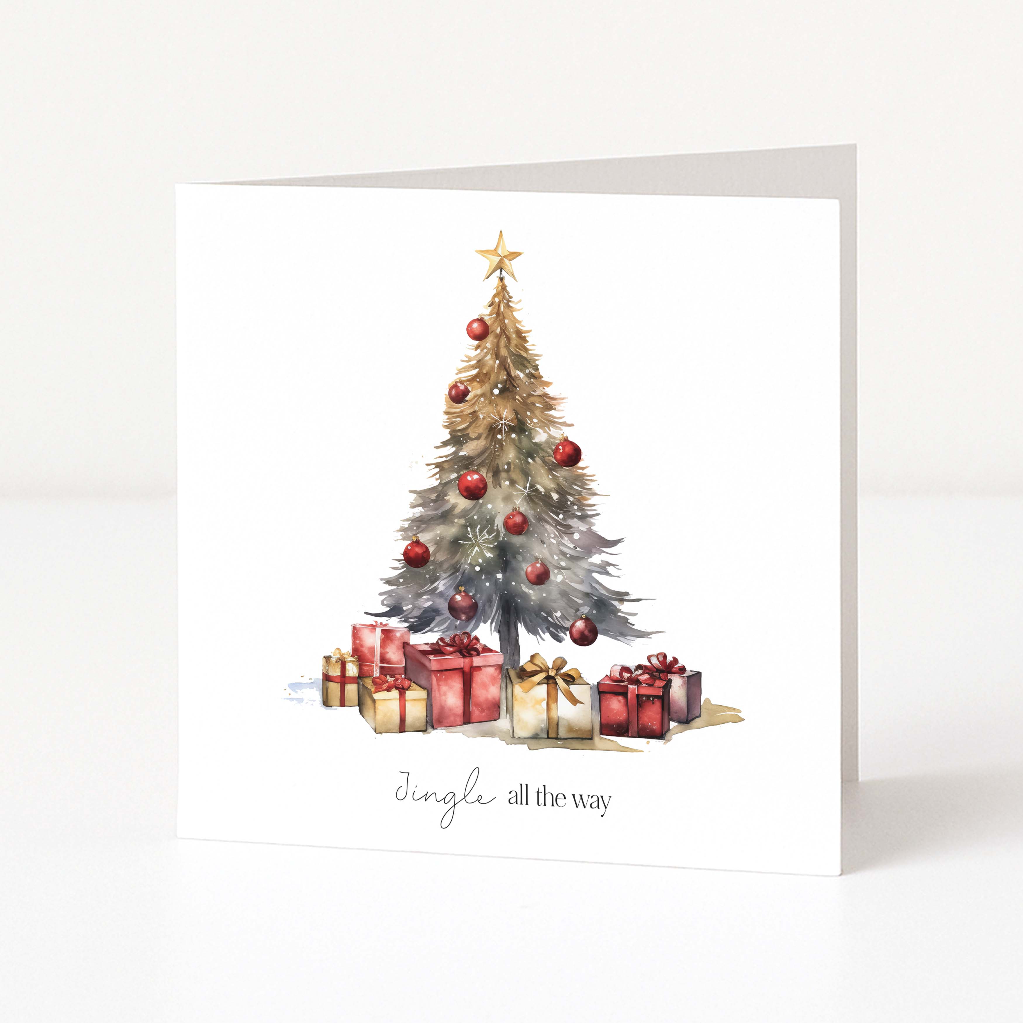 A white square Christmas greeting card standing upright, printed with a watercolour Christmas tree decorated with red ornaments and wrapped presents beneath a gold star, with the phrase 'Jingle all the way' at the bottom.