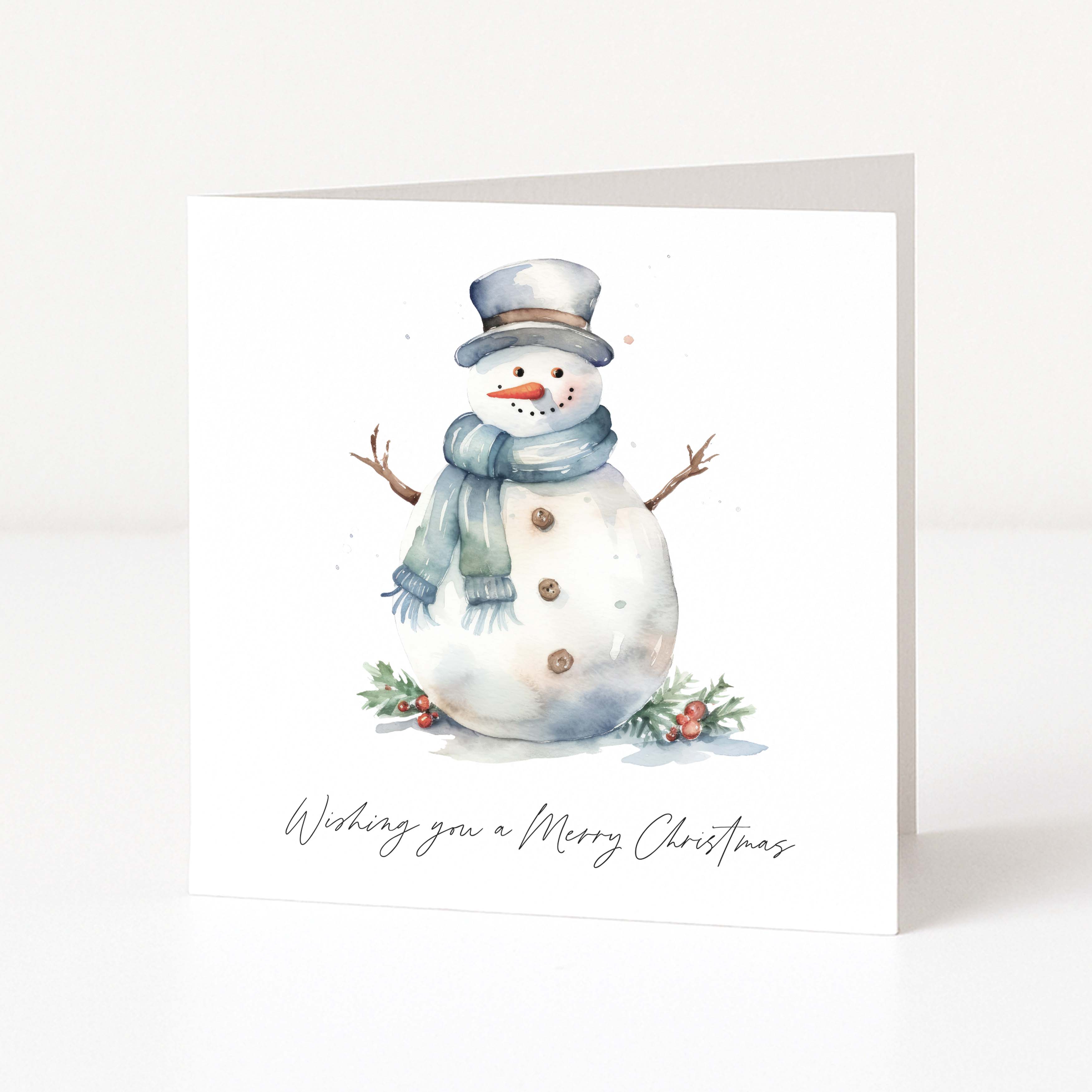 A square white Christmas greeting card standing upright, printed with a watercolor snowman wearing a gray top hat and blue scarf, with small greenery and red berries at the base and the text 'Wishing you a Merry Christmas' below.
