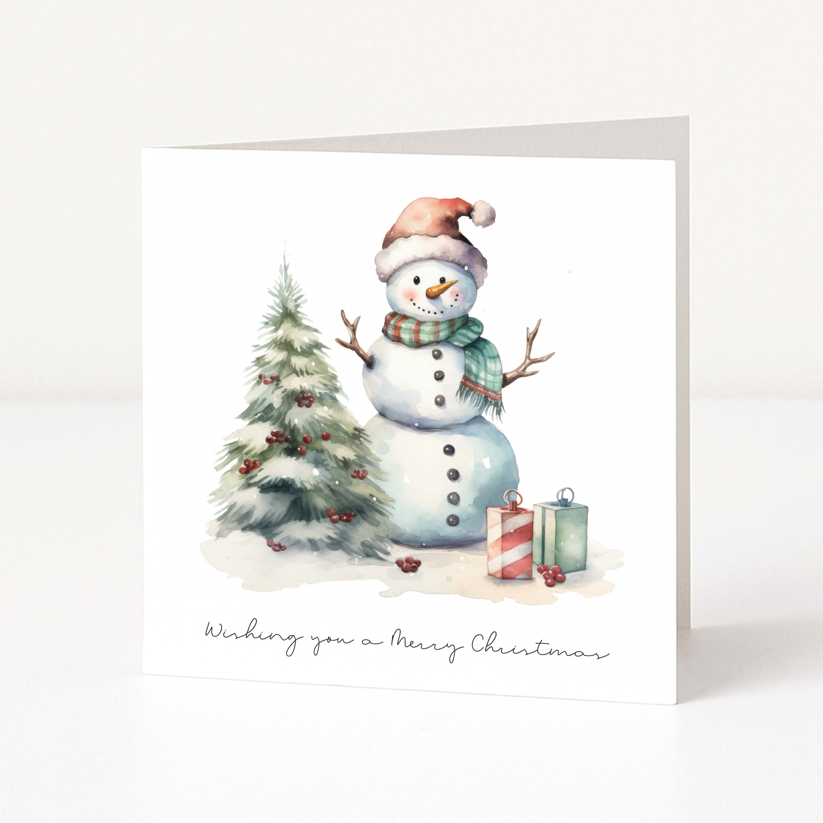 A square Christmas greeting card standing upright on a white surface. The front shows an illustrated snowman wearing a Santa hat and plaid scarf beside a small decorated tree and wrapped gifts, with the text 'Wishing you a Merry Christmas' at the bottom.