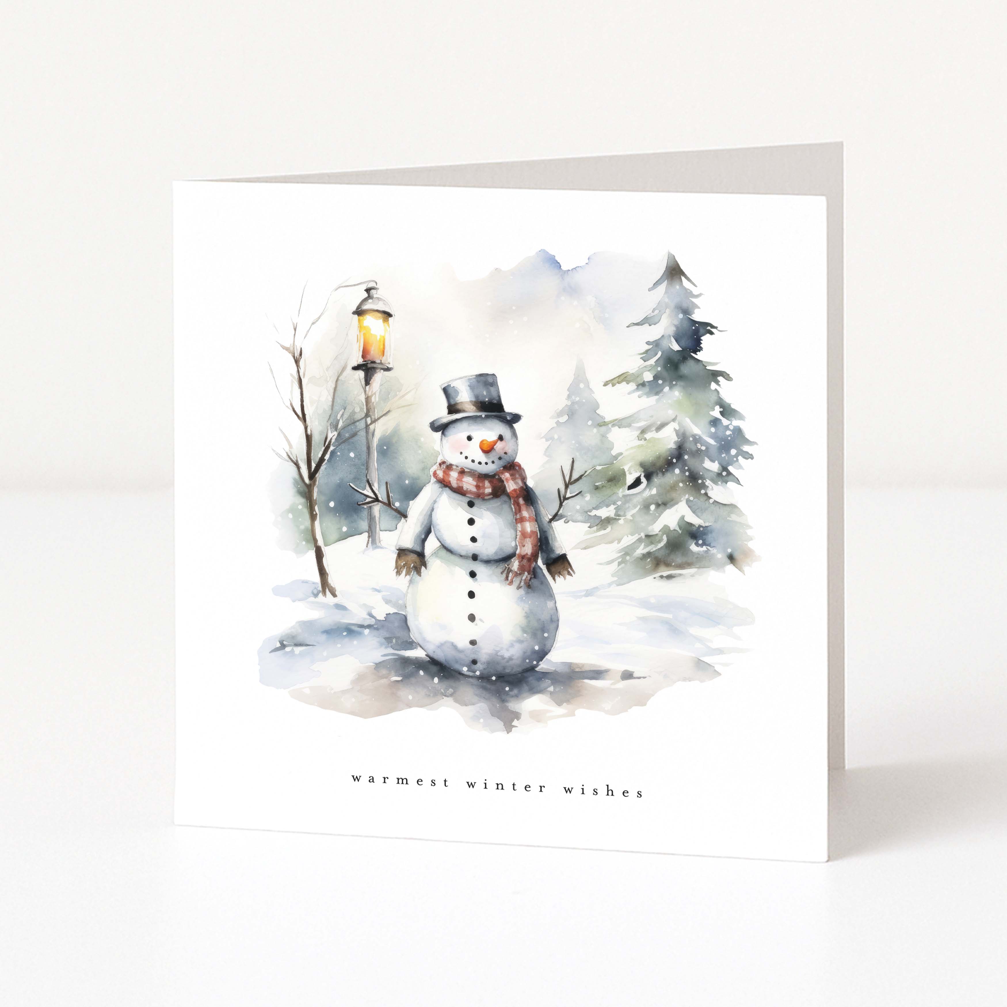 A white Christmas greeting card featuring a watercolour winter illustration of a snowman wearing a black top hat and a red-and-green plaid scarf, standing in a snowy landscape with bare trees, evergreen trees, and a glowing street lamp. The text 'warmest winter wishes' is printed below the illustration.