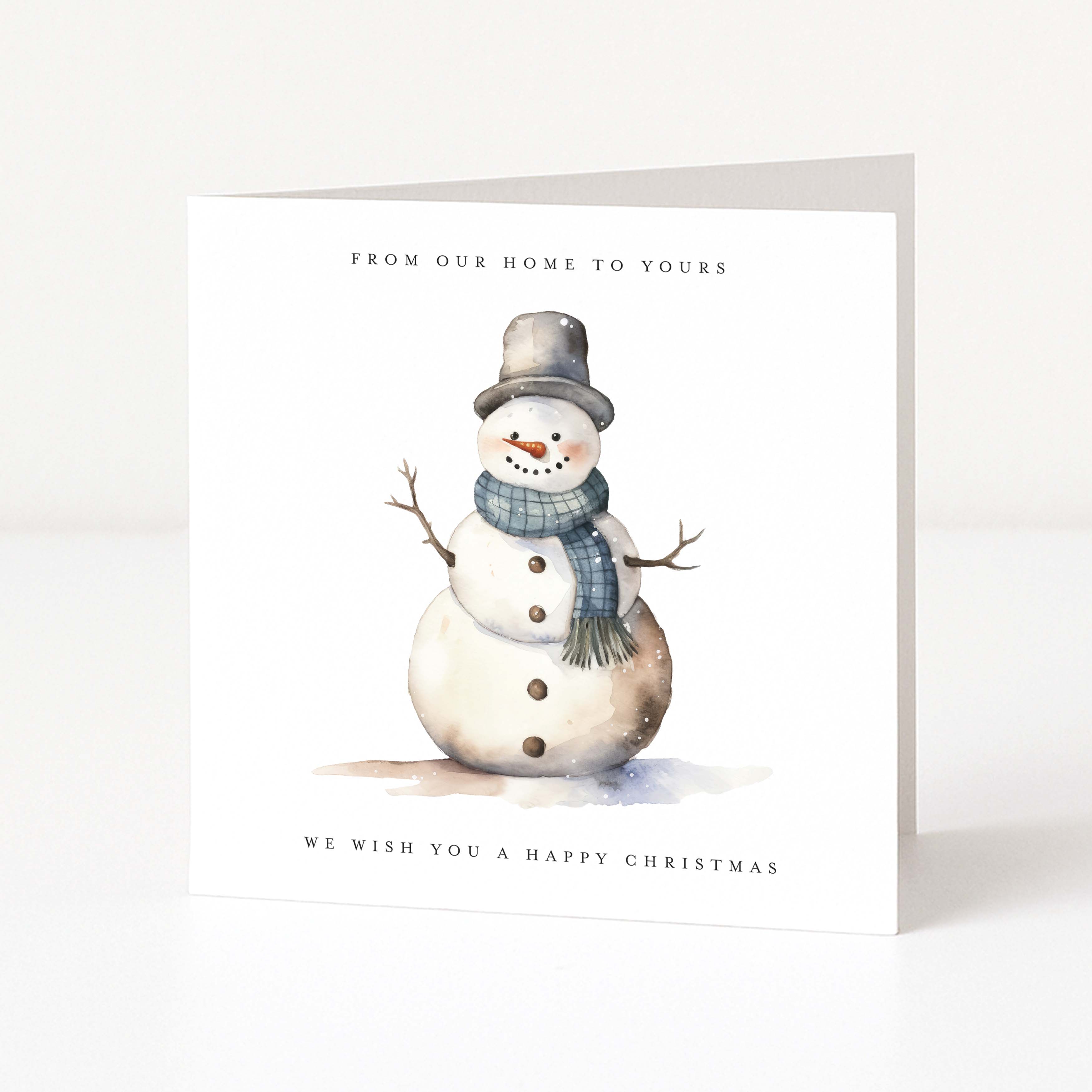 A square white Christmas greeting card standing upright on a light gray surface. The front features a watercolor-style snowman wearing a gray top hat and blue plaid scarf, with the text 'FROM OUR HOME TO YOURS' above and 'WE WISH YOU A HAPPY CHRISTMAS' below.