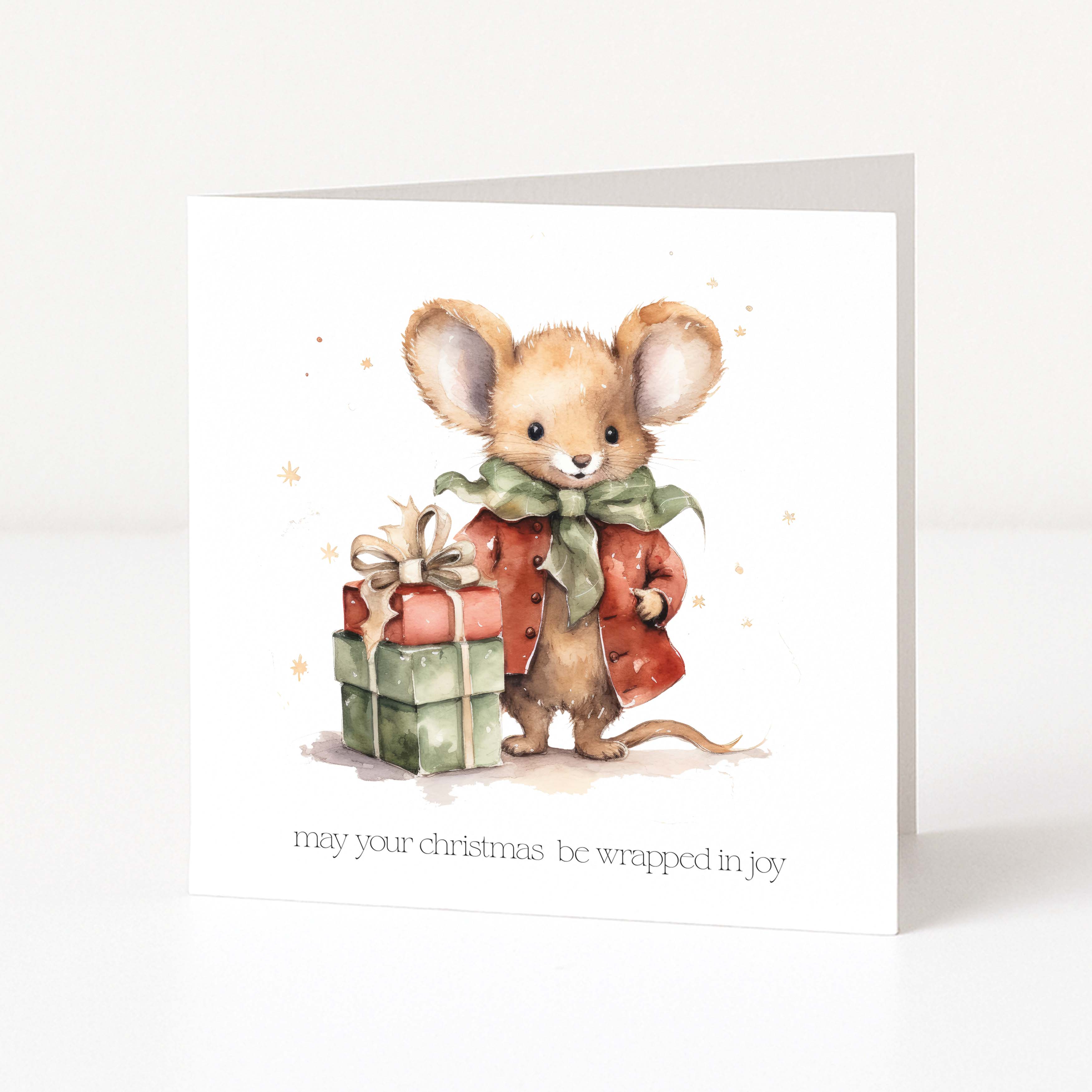 A square Christmas greeting card shown standing upright on a white surface. The card features a watercolour illustration of a small brown mouse wearing a red coat and green bow, standing next to a wrapped gift box.