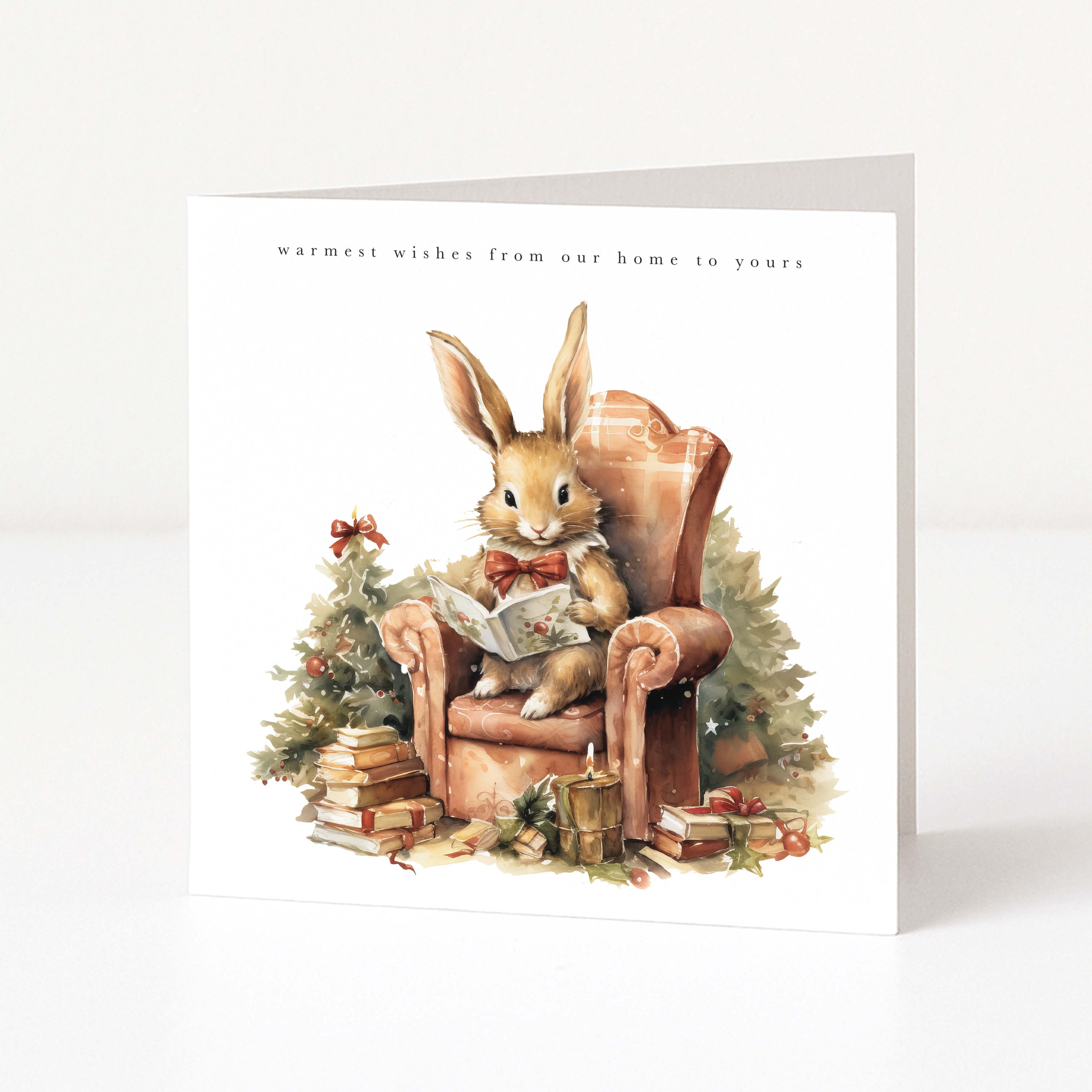 A square greeting card standing upright on a white surface against a light grey background. The front of the card features a watercolour-style illustration of a rabbit sitting in a brown armchair and reading a book, with a small decorated tree, books, and gifts around it.