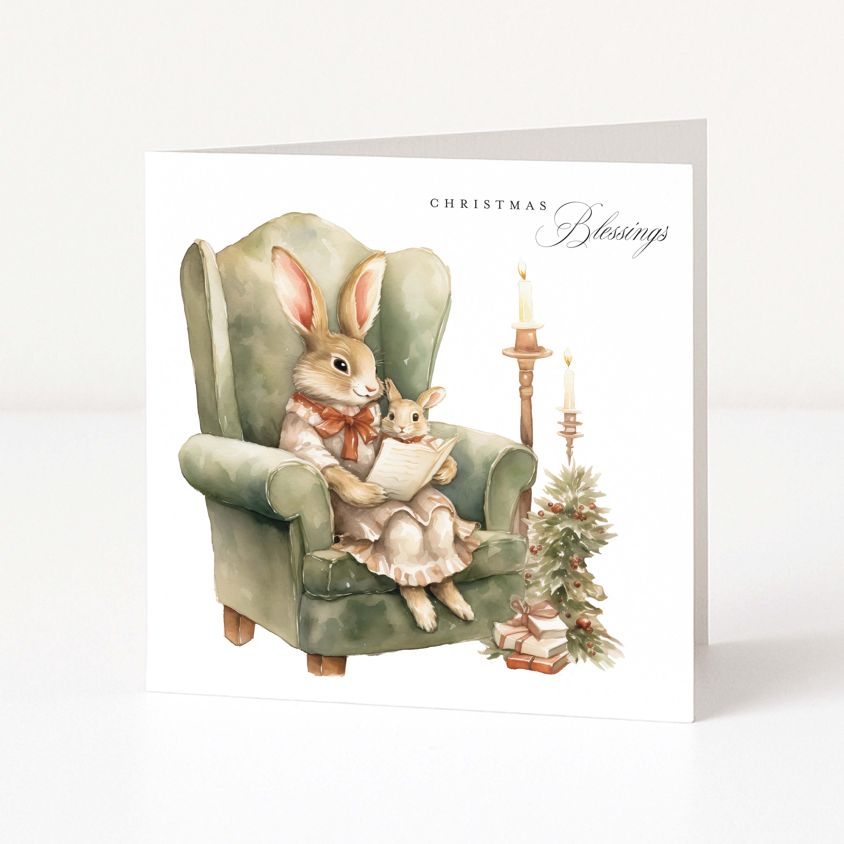 A square Christmas greeting card standing upright on a white surface. The front illustration shows a watercolour-style scene of two rabbits in a green armchair, with candles and a small decorated tree beside them.
